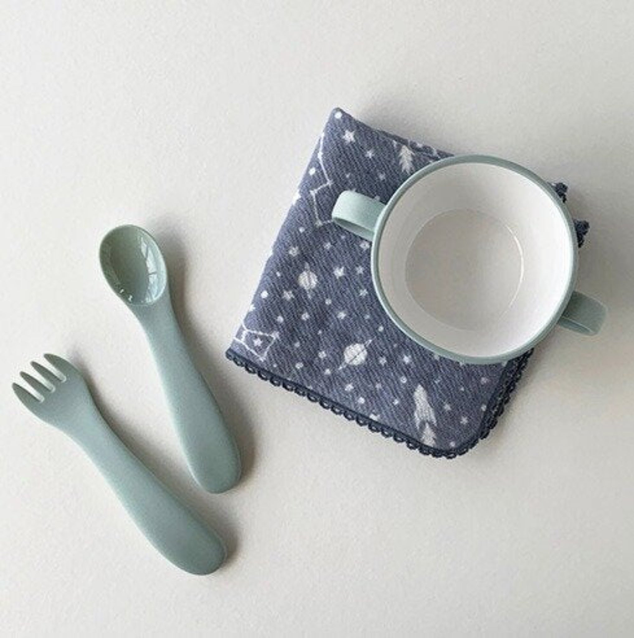 A folded blue face towel with a space print and delicate crotchet edge with a children's ceramic cup on top and a spoon, and fork beside.