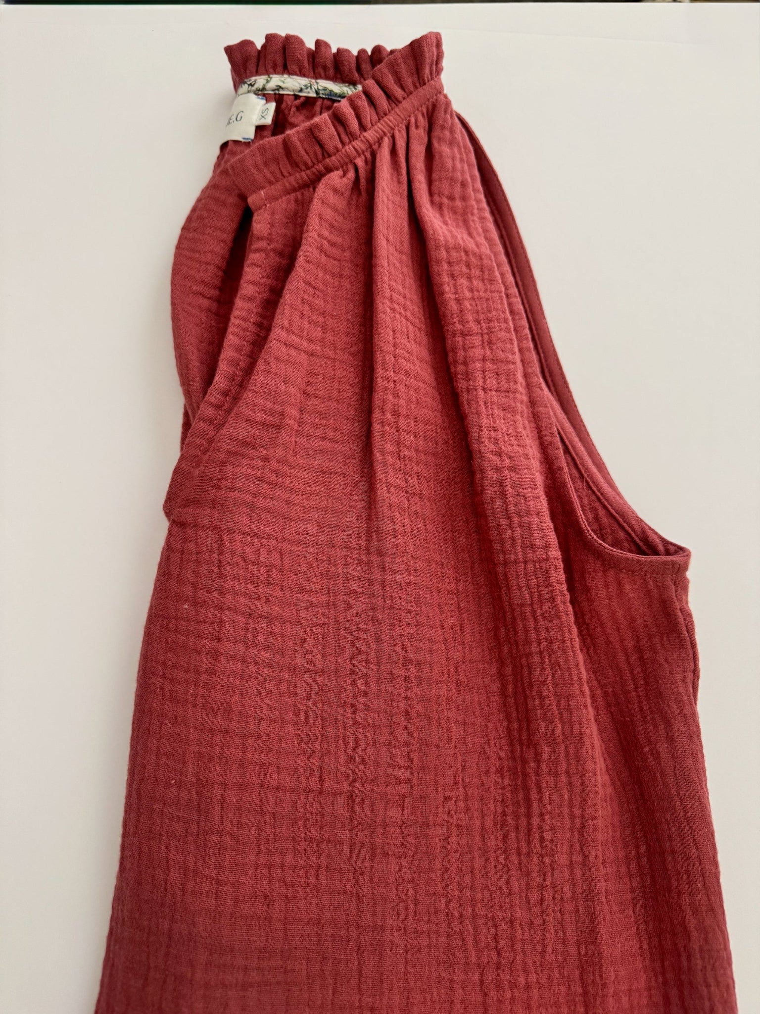 Maroon cotton gauze sleeveless top with frill around neck folded in half.