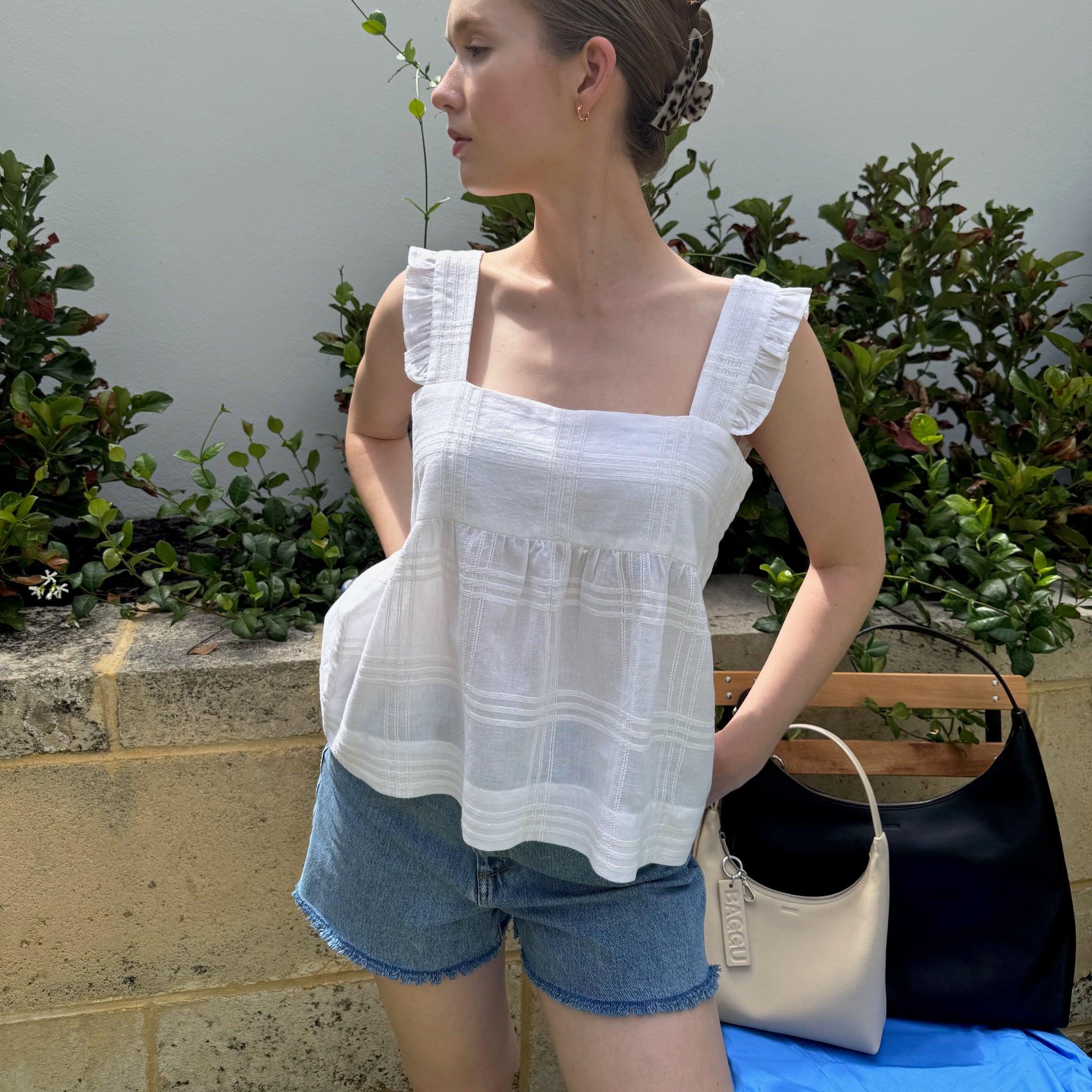 Woman wearing white cami top with frill strap and denim shorts in garden with leather bags on chair.