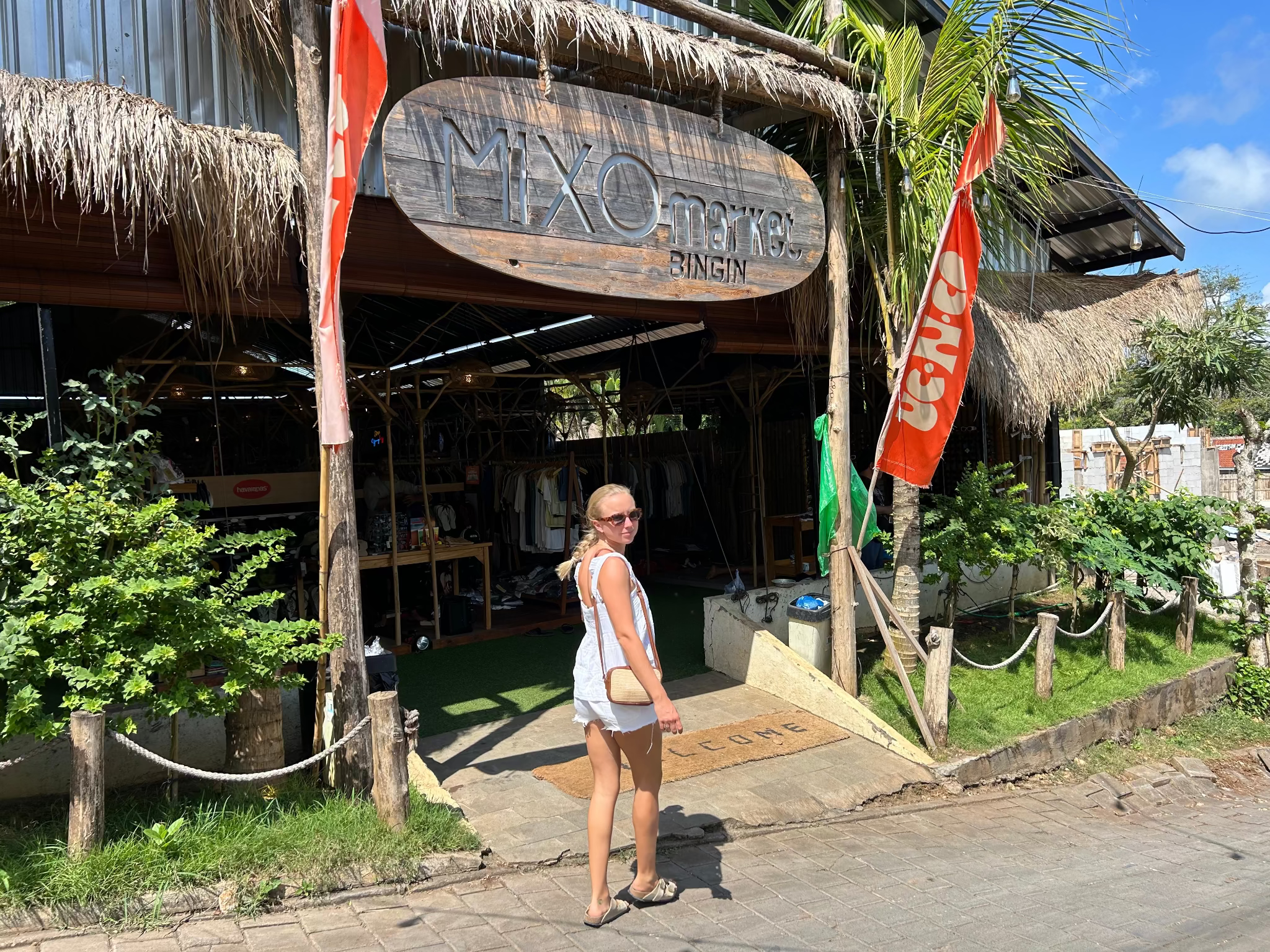 Woman wearig white cami top and shorts standing outside a market with a sign that reads 'Mix' on a sunny day.