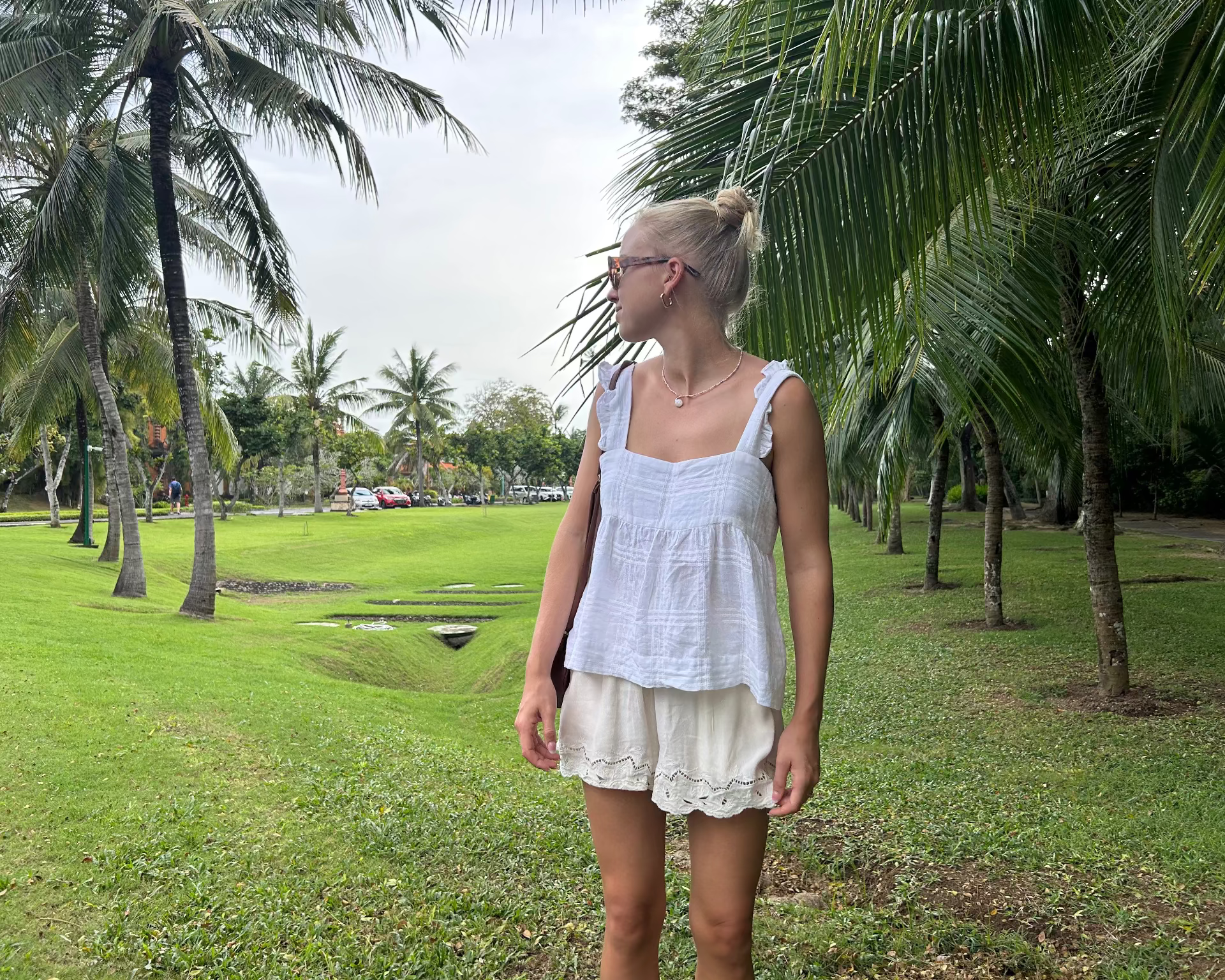 Woman in a white cami top and beige shorts standing in a park with palm trees.
