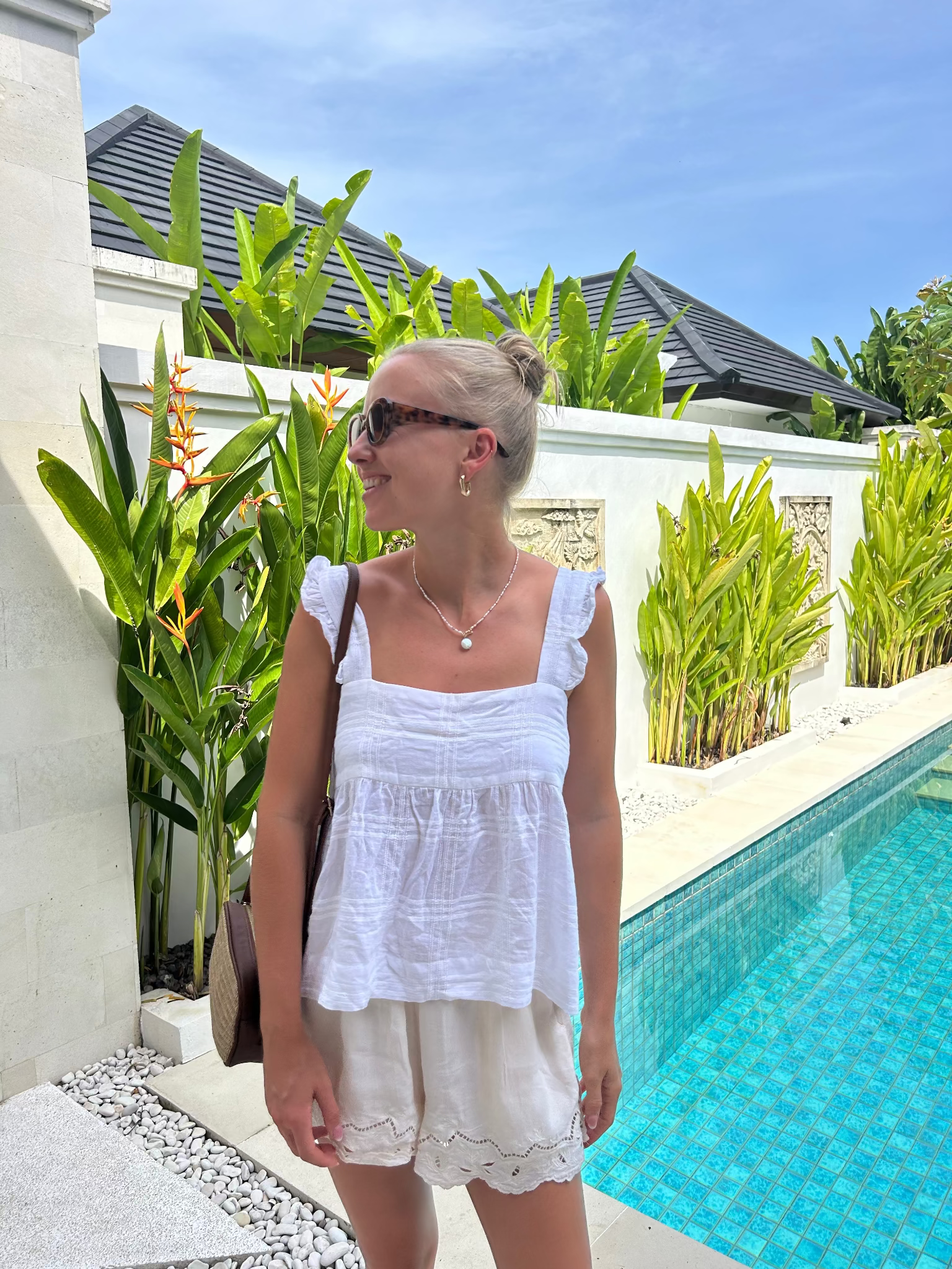 Woman in a white cami top with frill on strap and cream shorts outfit standing by a pool with greenery.