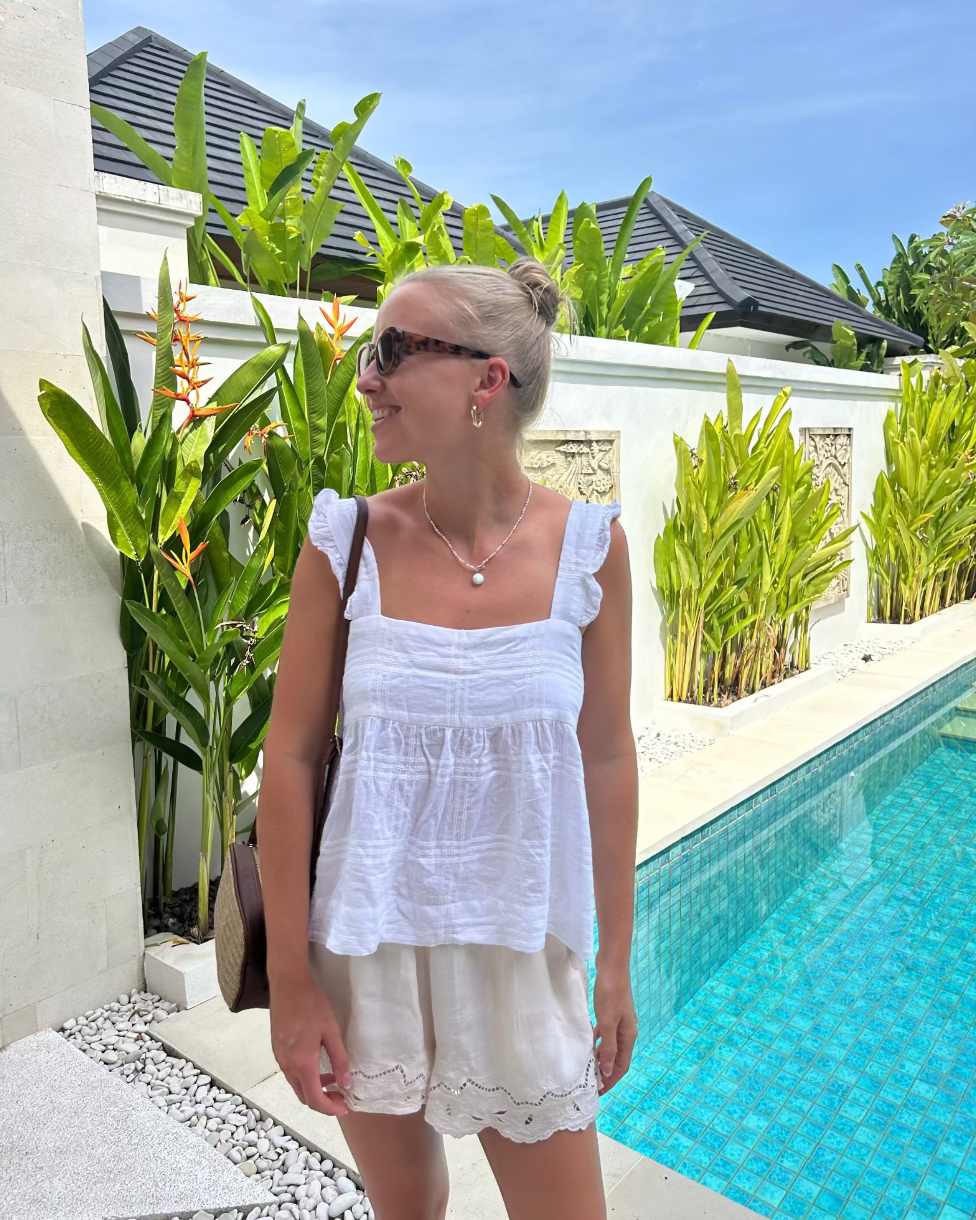 Woman in a white cami top with frill on strap and cream shorts outfit standing by a pool with greenery.