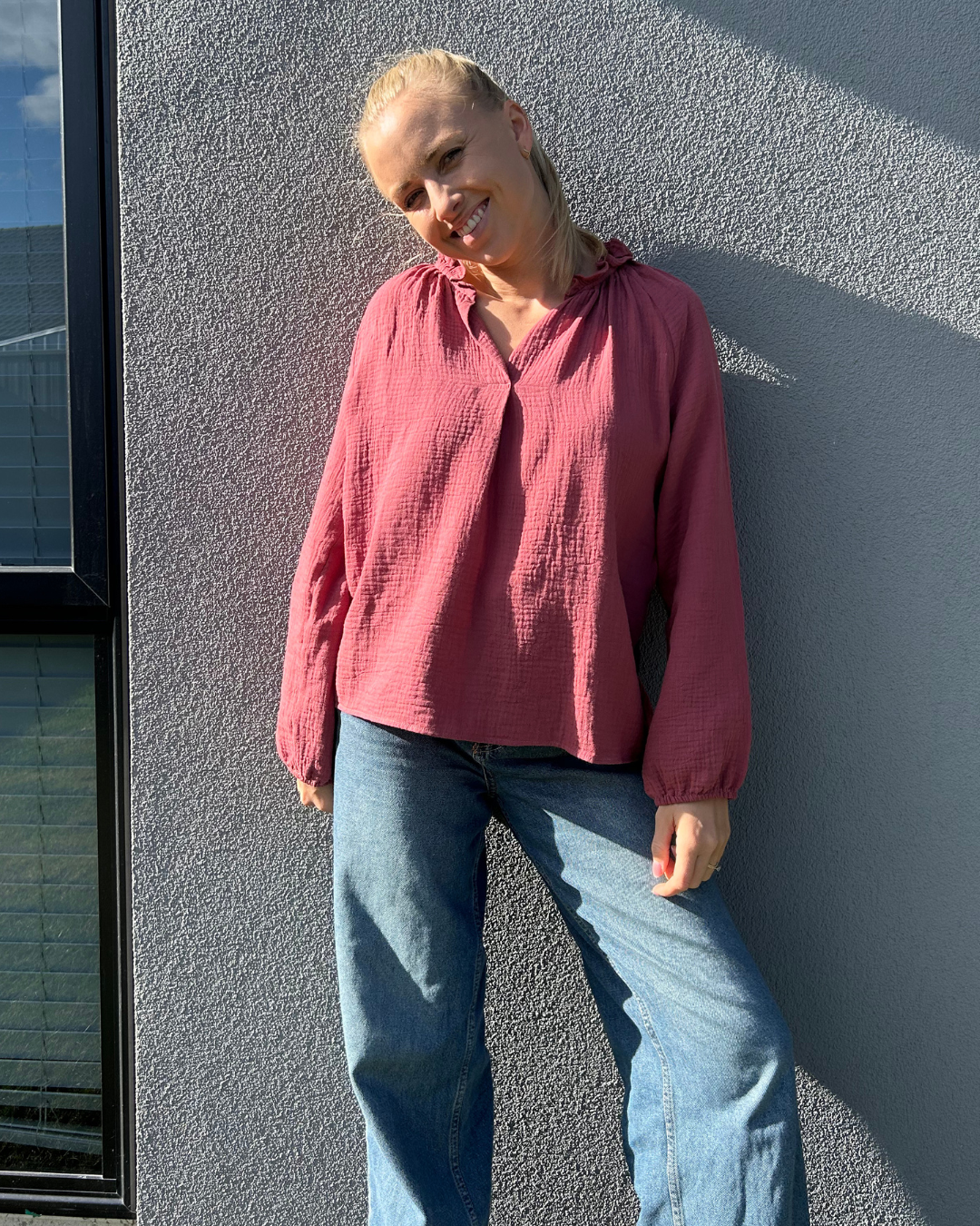 Person wearing a maroon blouse and blue jeans leaning against a grey wall.