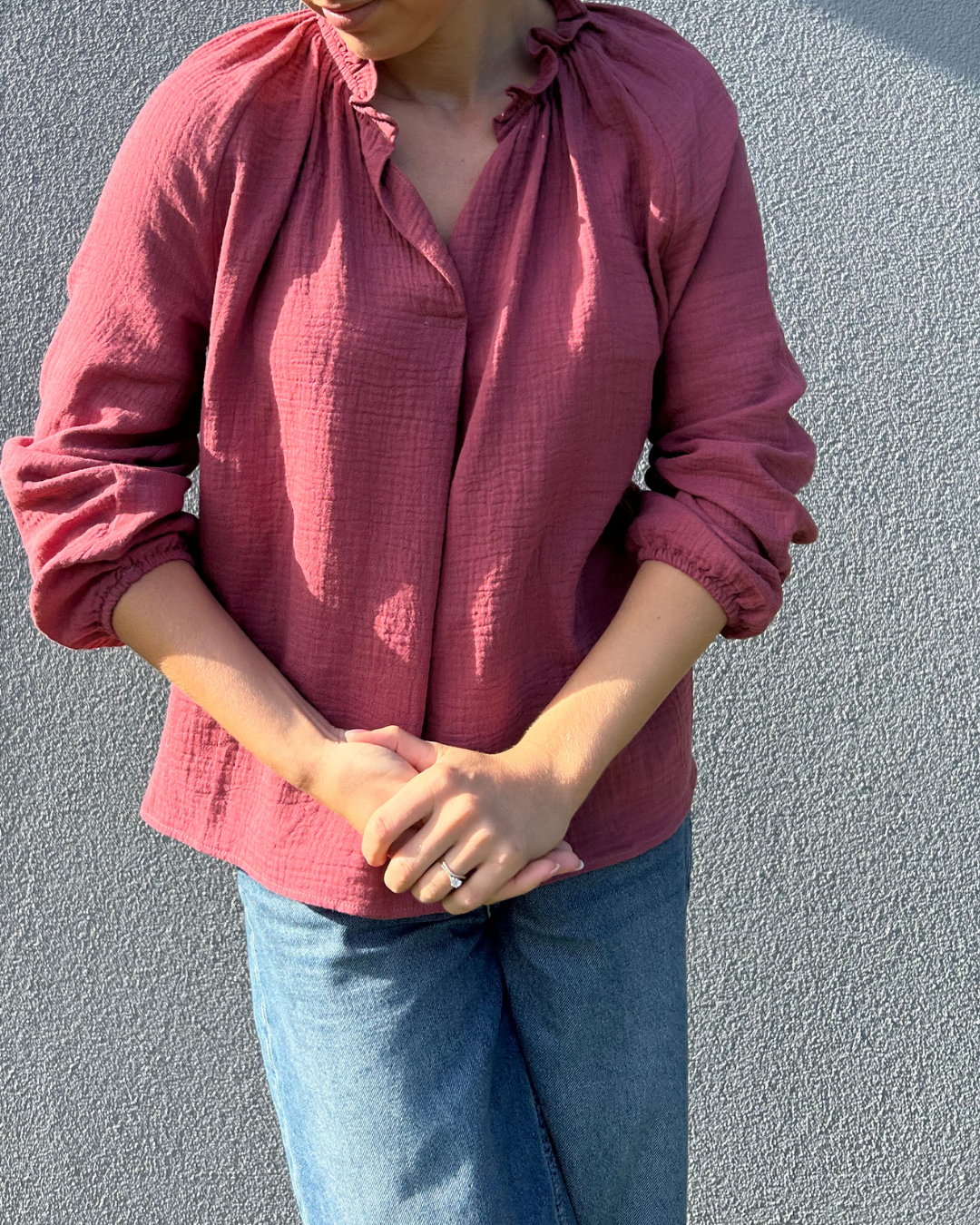 Person wearing a burgundy blouse with long sleeves and blue jeans against a grey wall.