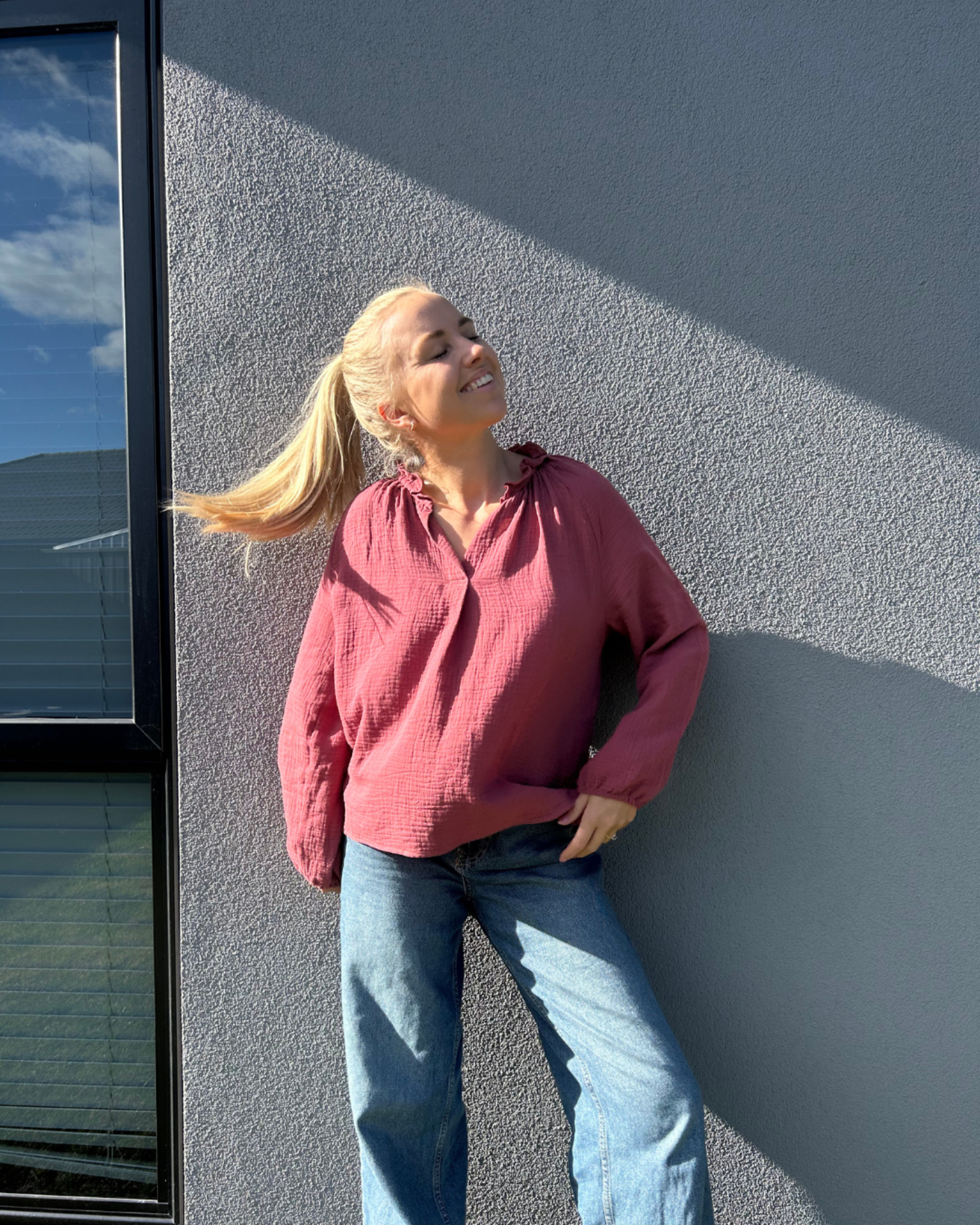 Woman in a maroon long sleeve shirt and blue jeans leaning against a gray wall.