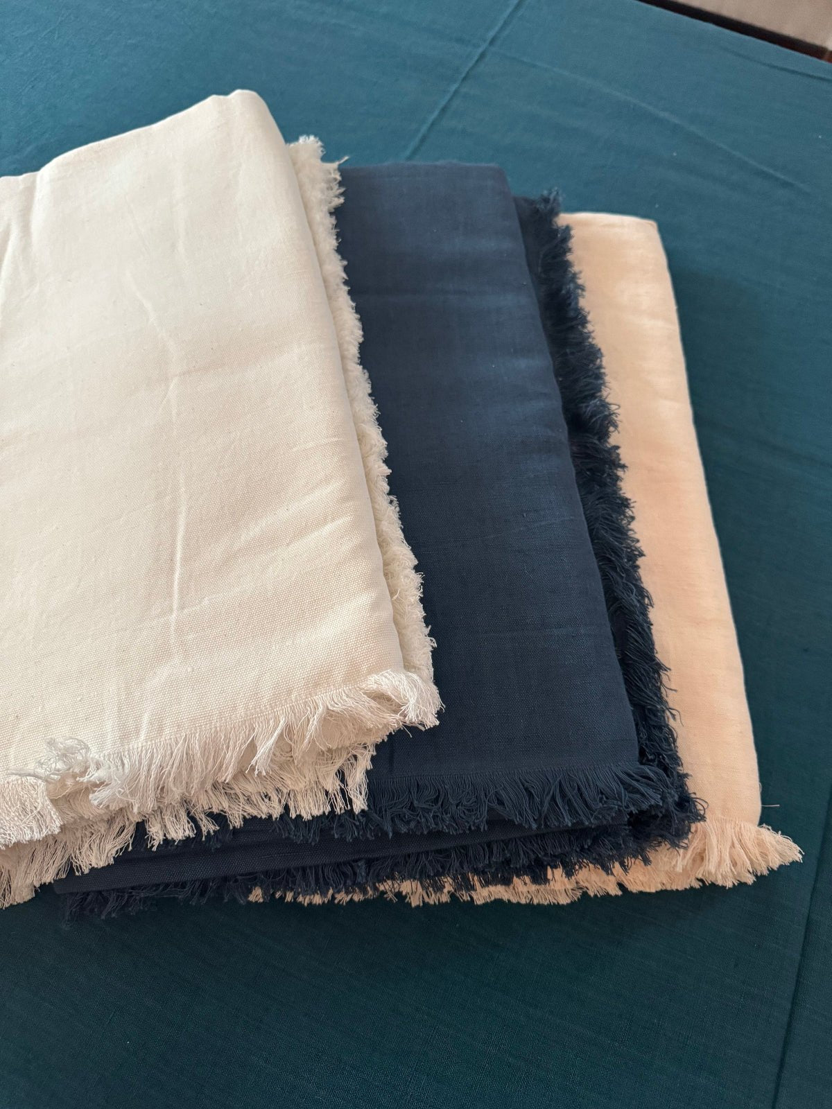 Four tablecloths in teal on the table with a stack of blush, ocean blue, and white with a fringe edge