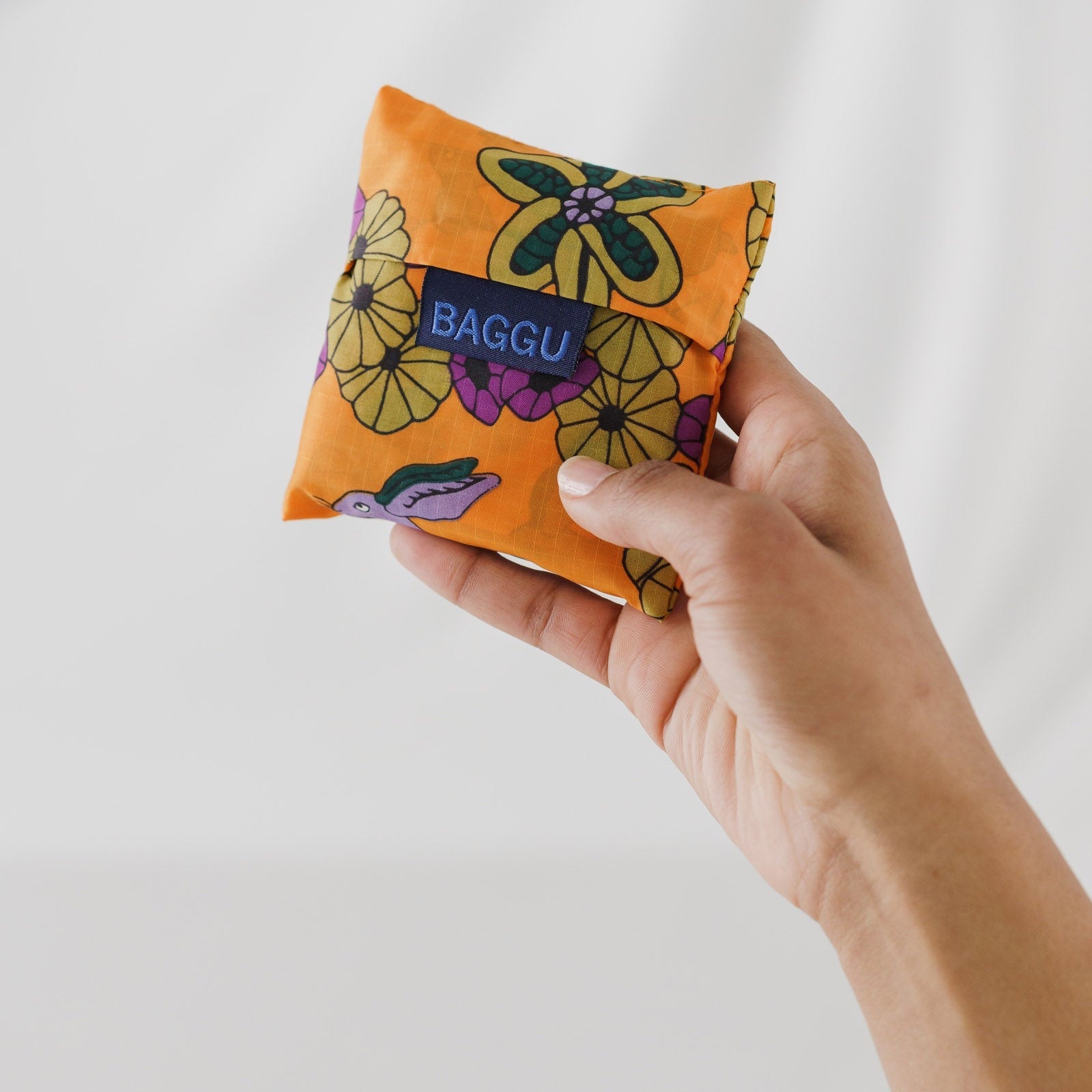 Hand holding an orange floral and purple rabbit baby size reusable bag storage bag with 'Baggu' branding