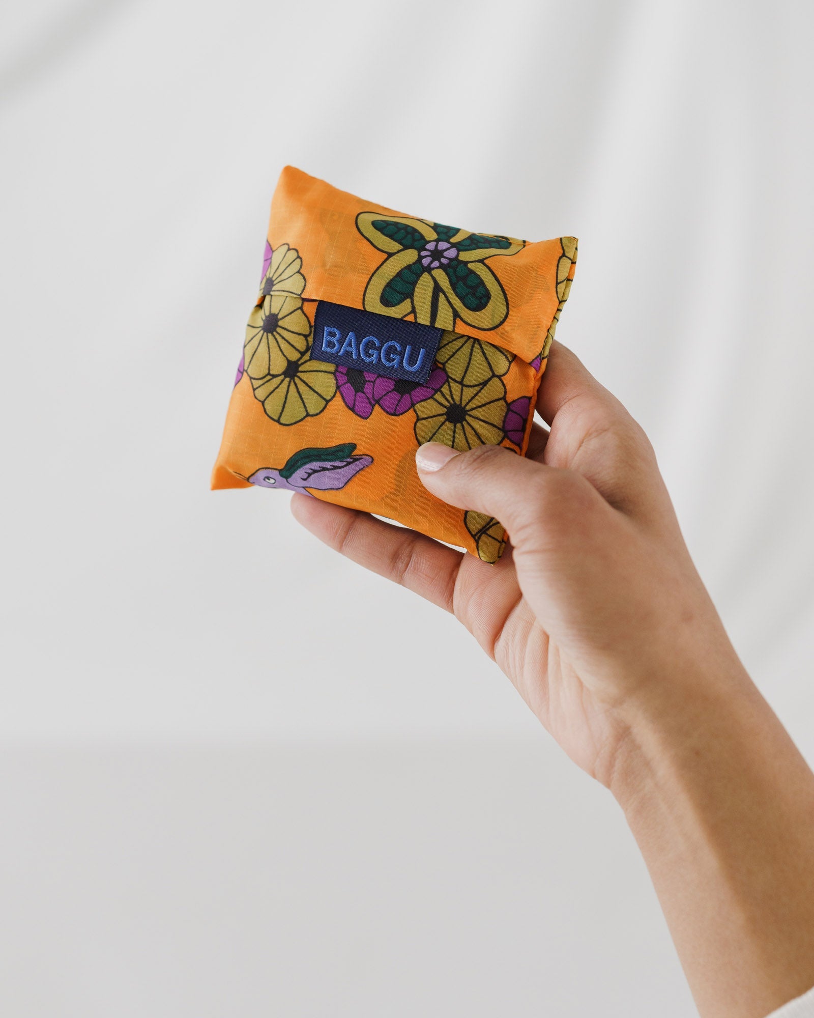 Hand holding an orange floral and purple rabbit baby size reusable bag storage bag with 'Baggu' branding