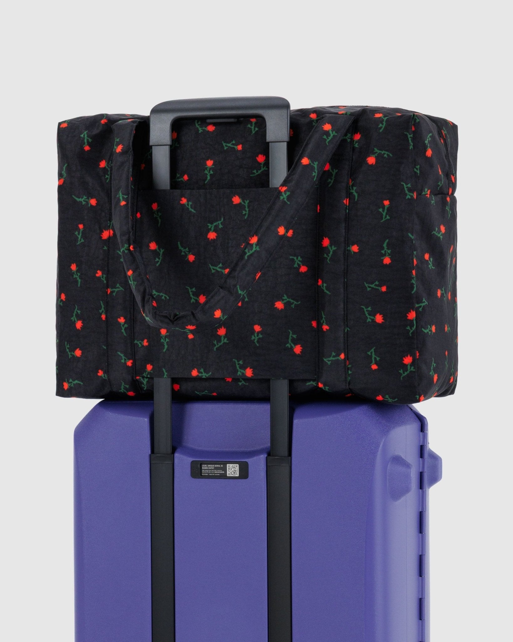 Large black duffle bag with red wildflowers pattern sitting on top a purple suitcase attached with travel sleeve at back
