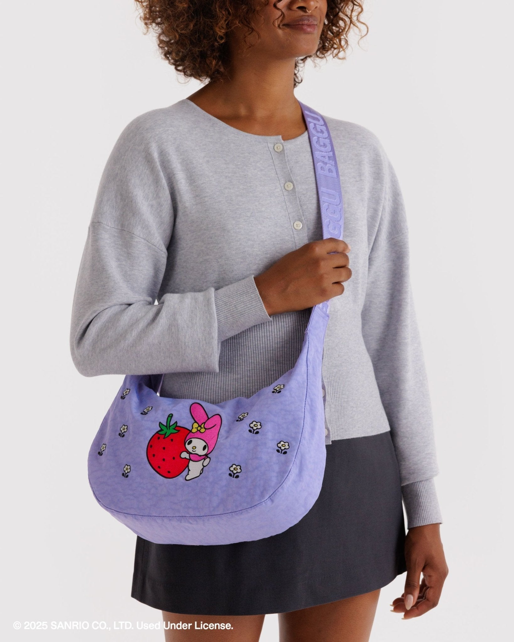 Person wearing a purple crescent shaped bag crossbody with hello kitty cartoon character holding a strawberry design and white small flowers on front