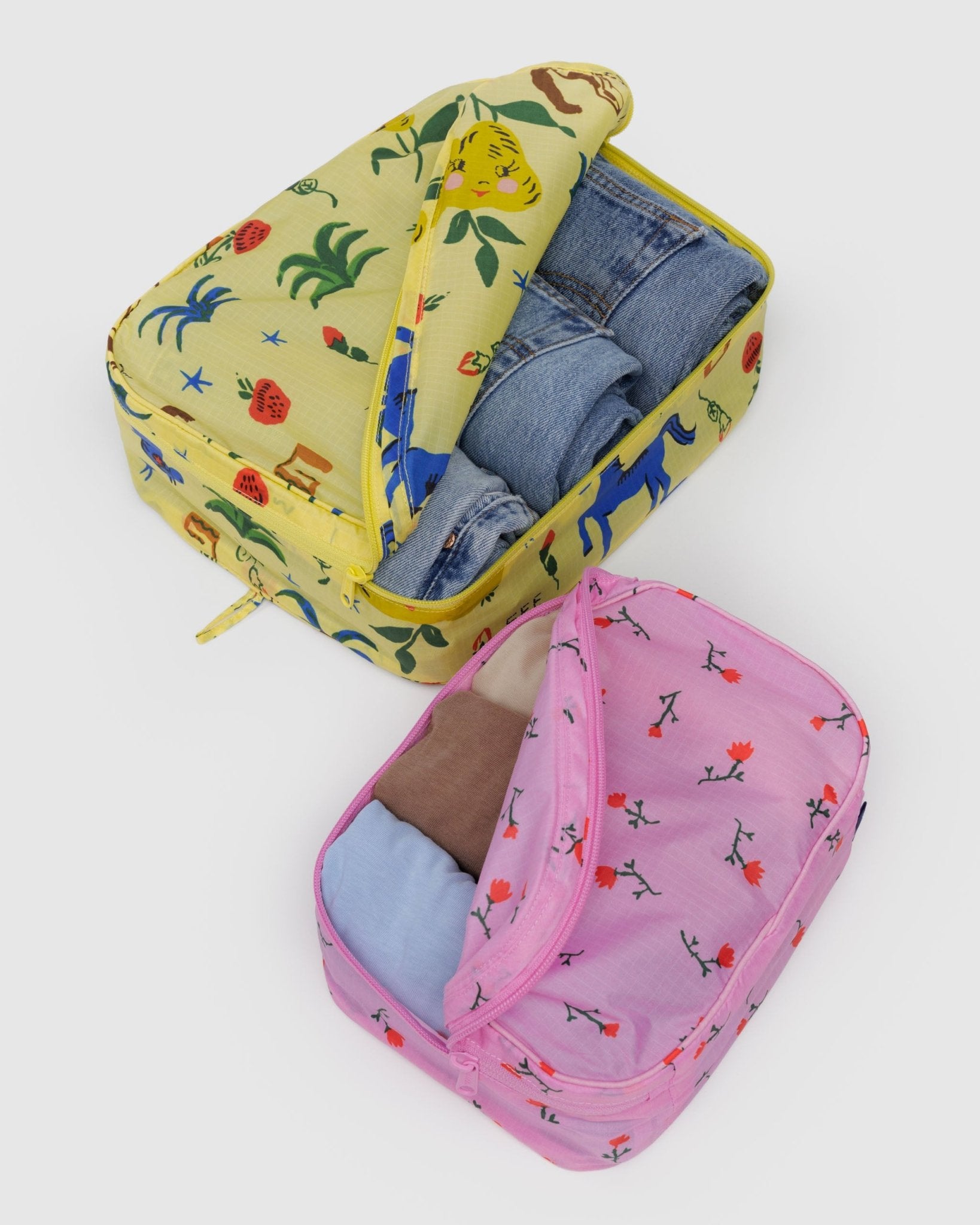 Two different sized packing cubes in yellow with horse and fruits print and pink with flowers filled with clothes inside
