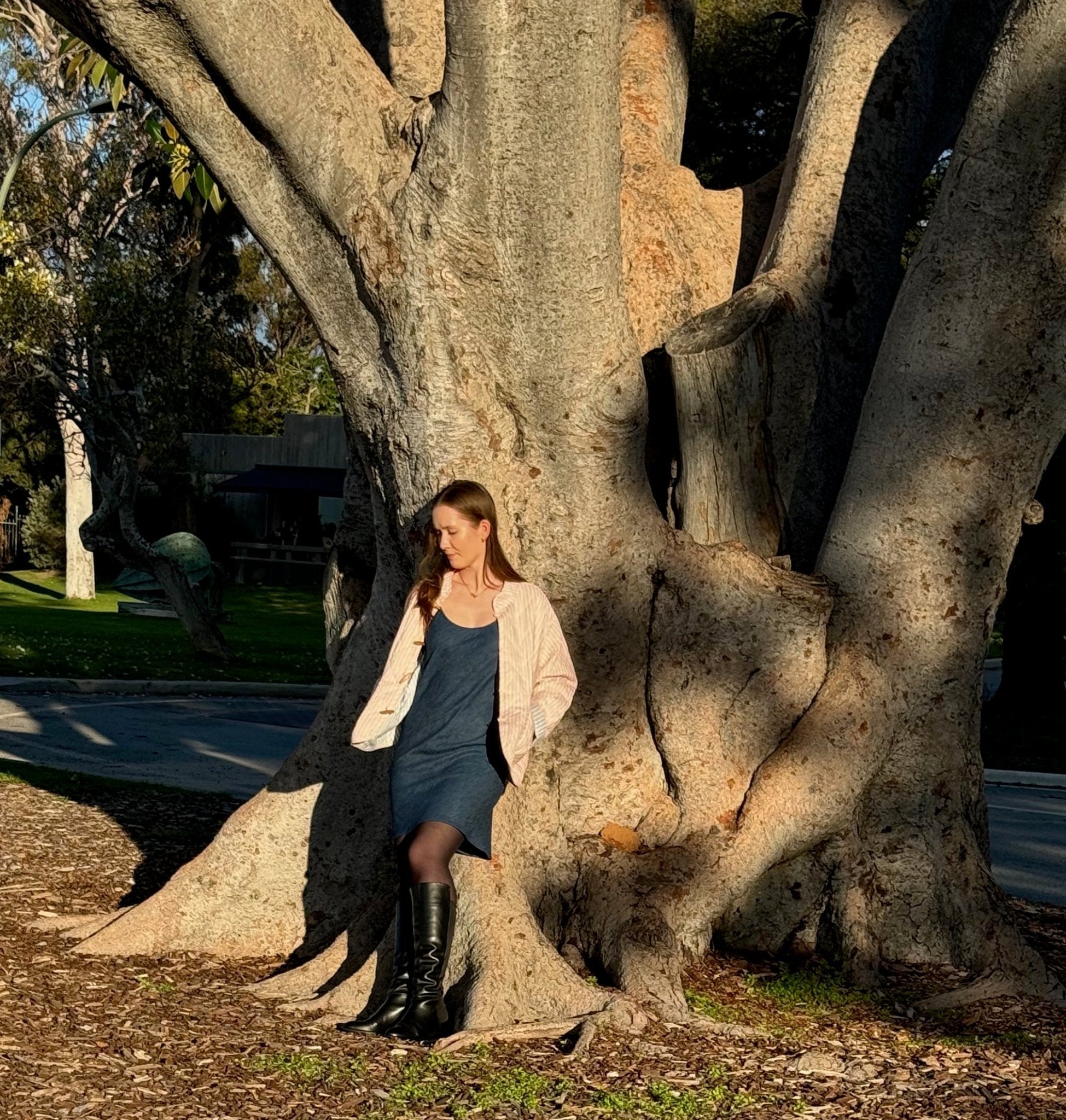 Woman wearing blue denim Bias Slip Mini Dress and pink stripe jacket  leaning on a tree.