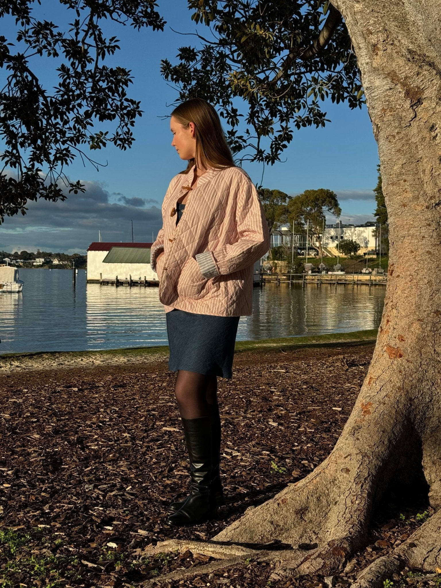 Side of a woman wearing a blue denim Slip Mini Dress and pink stripe quilted jacket near a tree and water with hands in pockets.