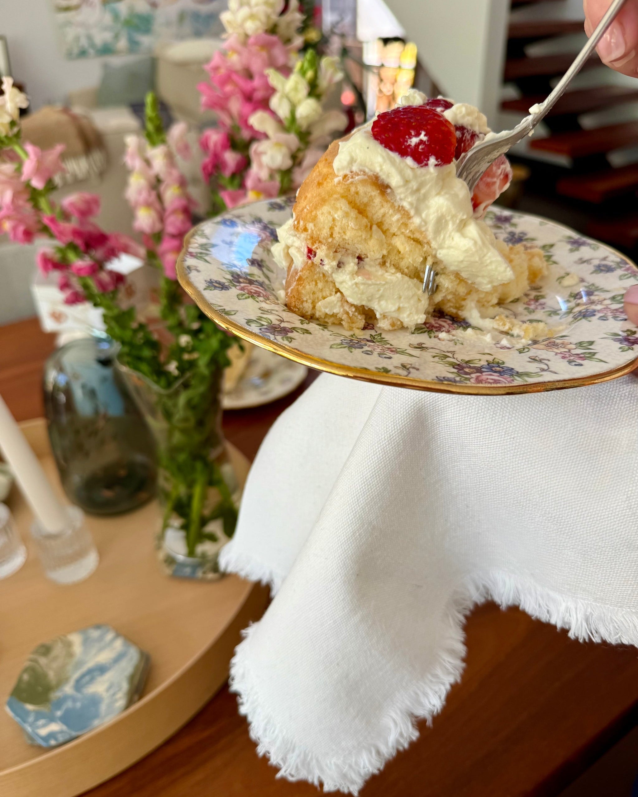 Persons hand holding a white napkin with frayed edge detail and a slice of cake on a decorative plate with flowers in background