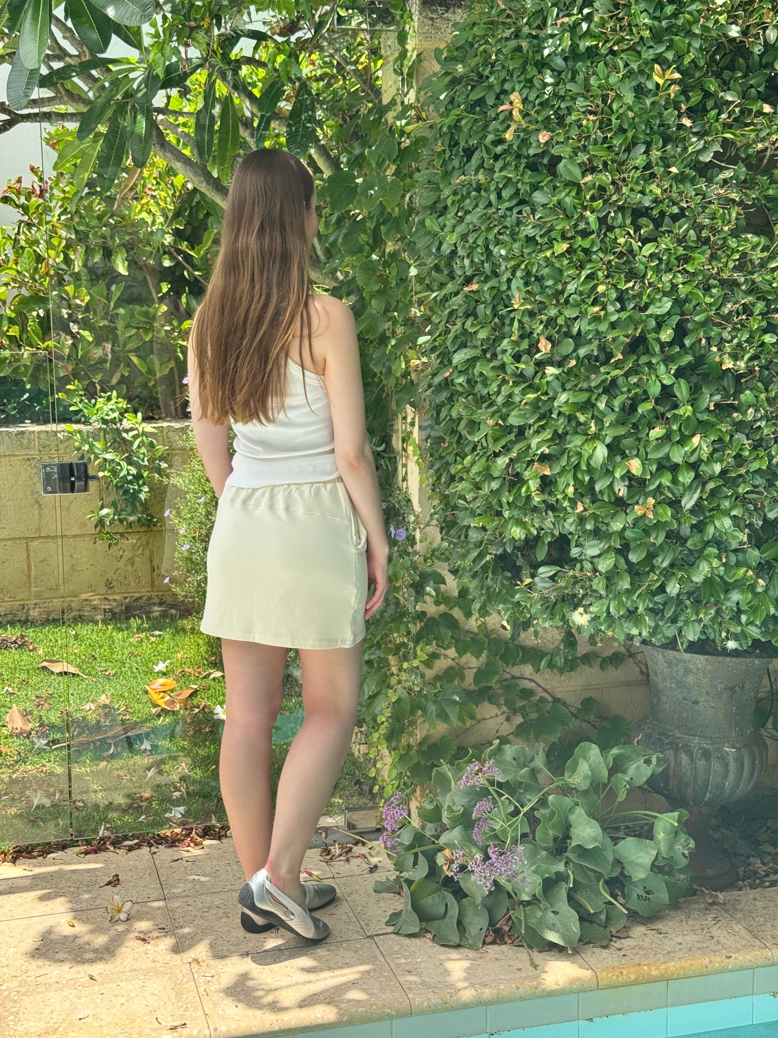 Woman wearing a light yellow miniskirt and white top standing in a garden with greenery and a pathway.