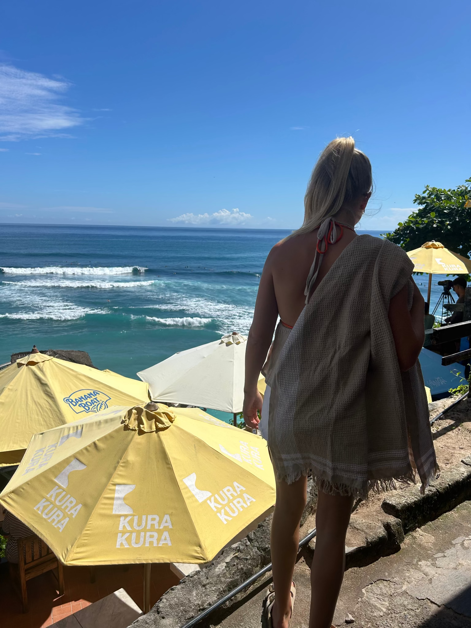 Person standing on a cliff overlooking the ocean with waffle towel on shoulder and yellow umbrellas.