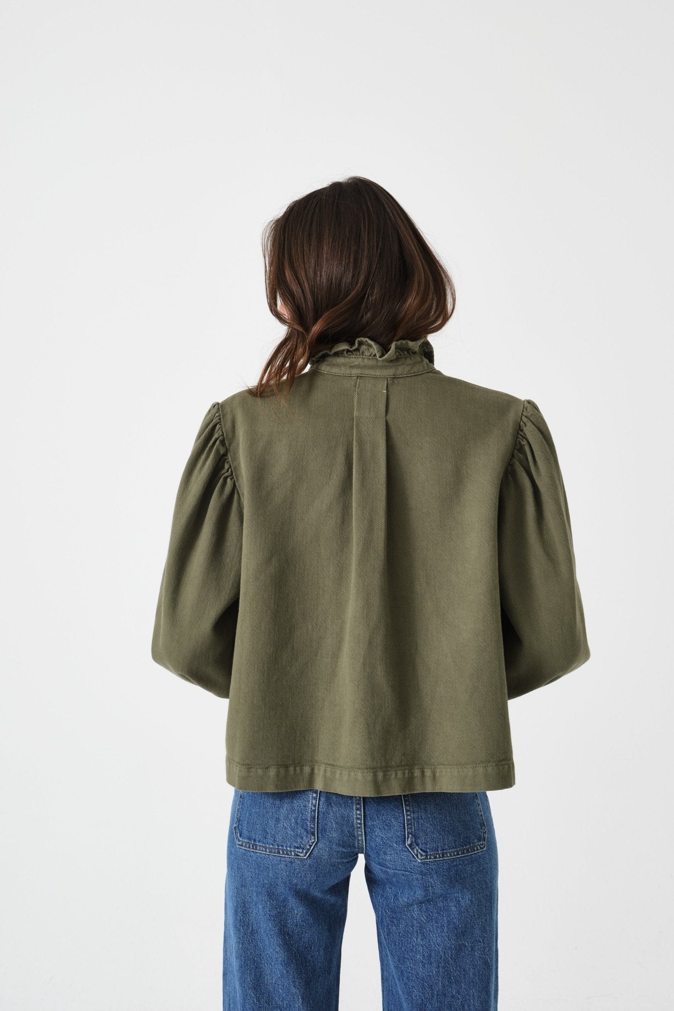 Back of woman wearing an olive green jacket with pleat at midback and frill collar, and blue jeans