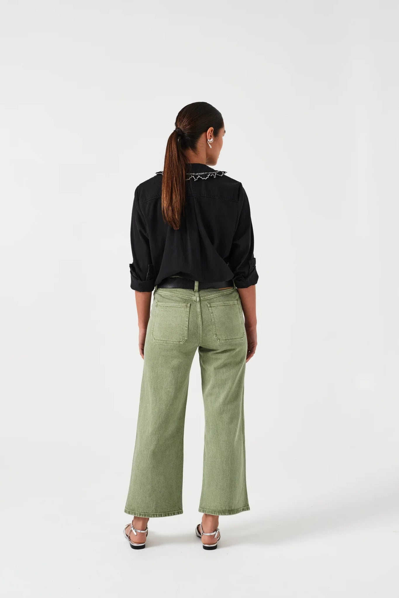 Woman facing backwards wearing green denim jeans with pockets a black shirt and sandals.