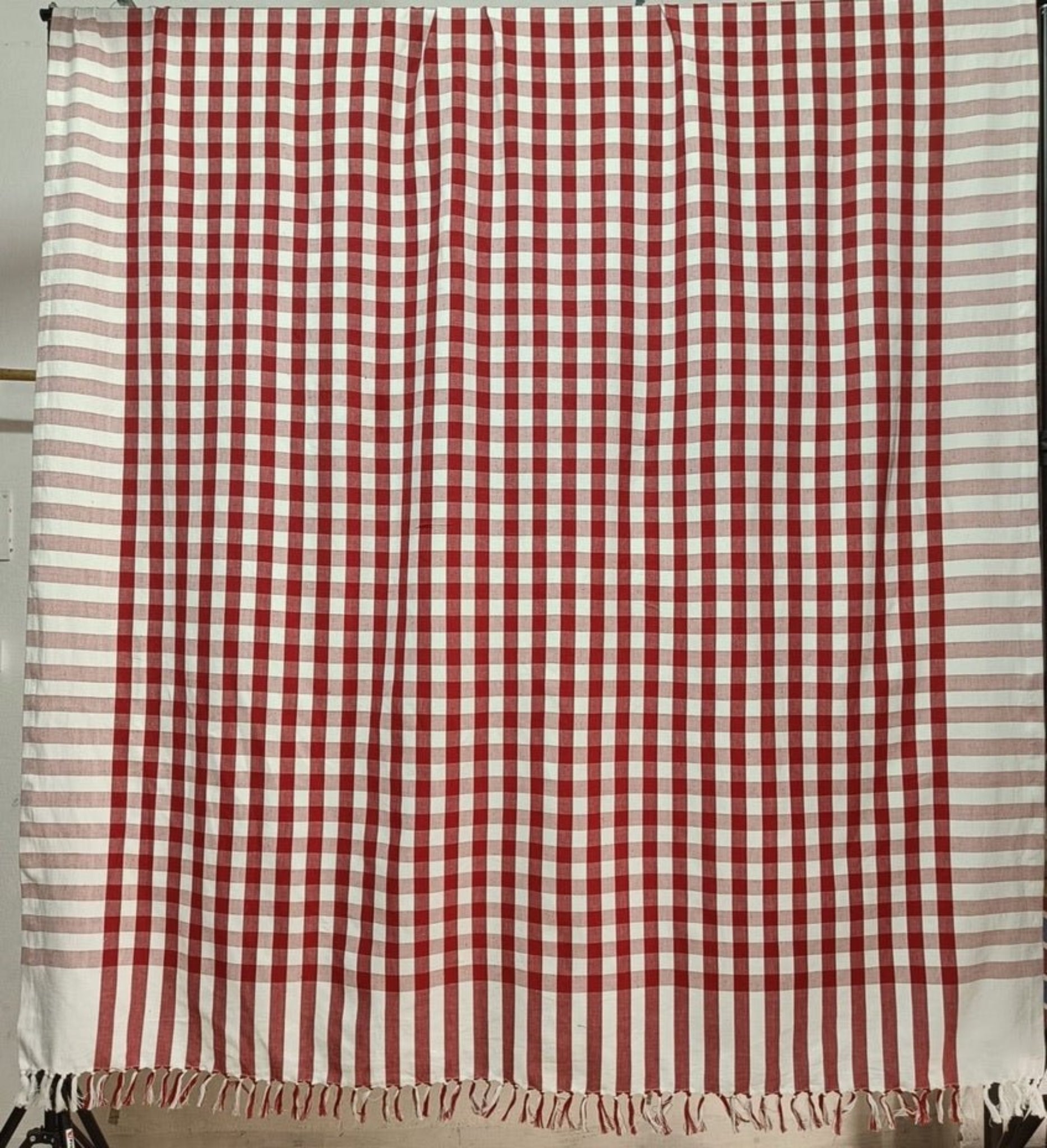 A hanging rectangular tablecloth in red check bordered by red and white stripe and a handknotted fringe.