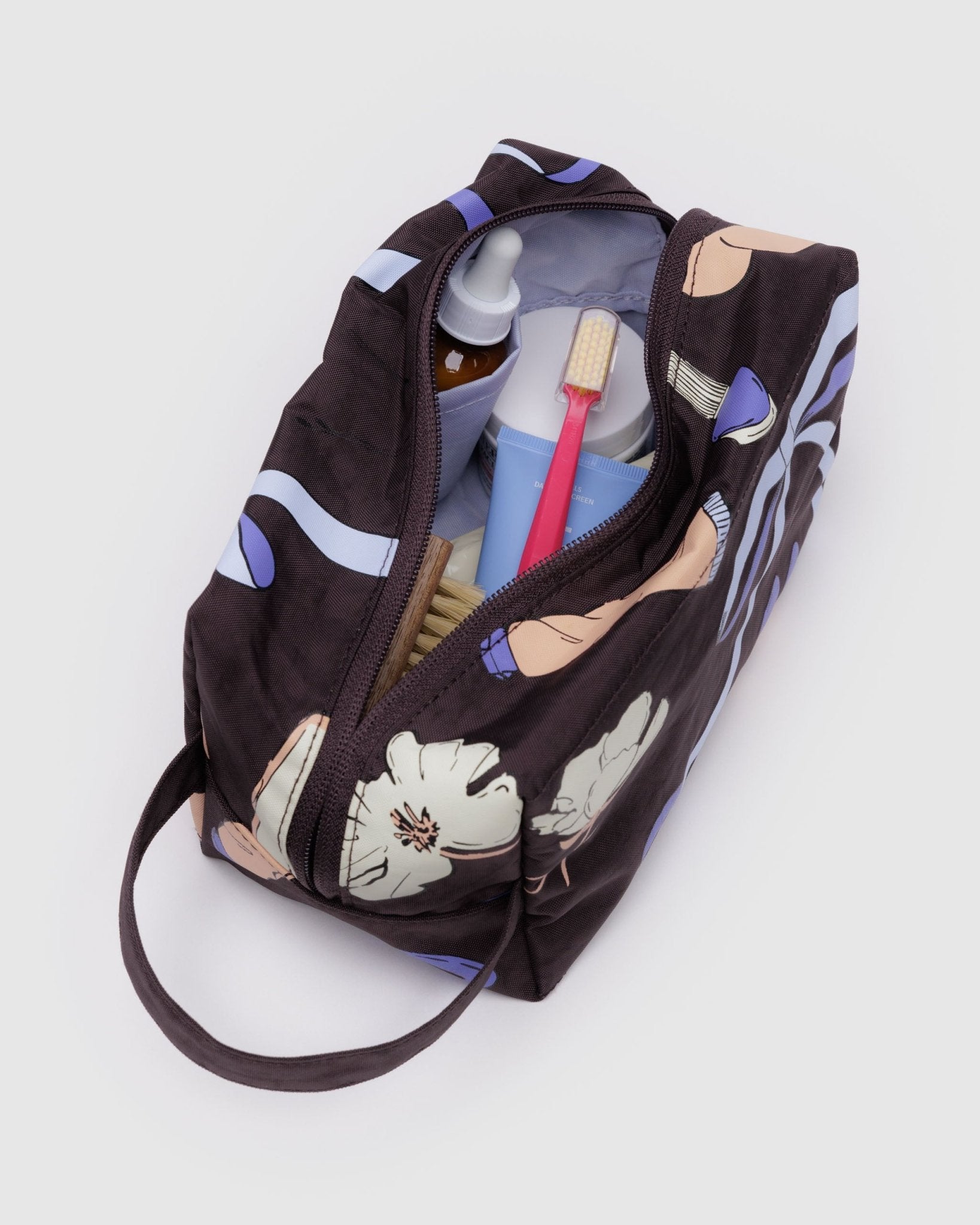 Carry handle on a brown dopp kit with prints of white flowers, purple bows, pink tube with wide zip top open, purple lining and toiletries inside.