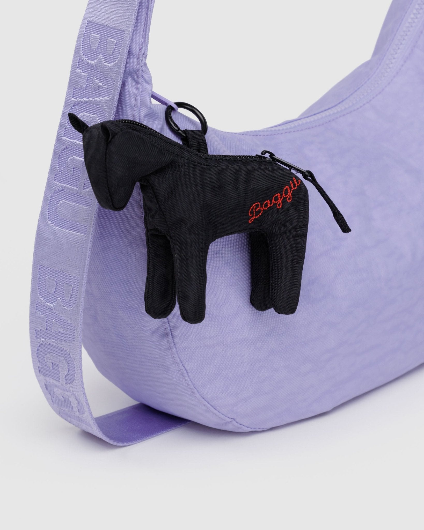 Baggu embroidered branding on side of black puffy Horse bag charm with zip along back attached to zip of purple crescent bag.