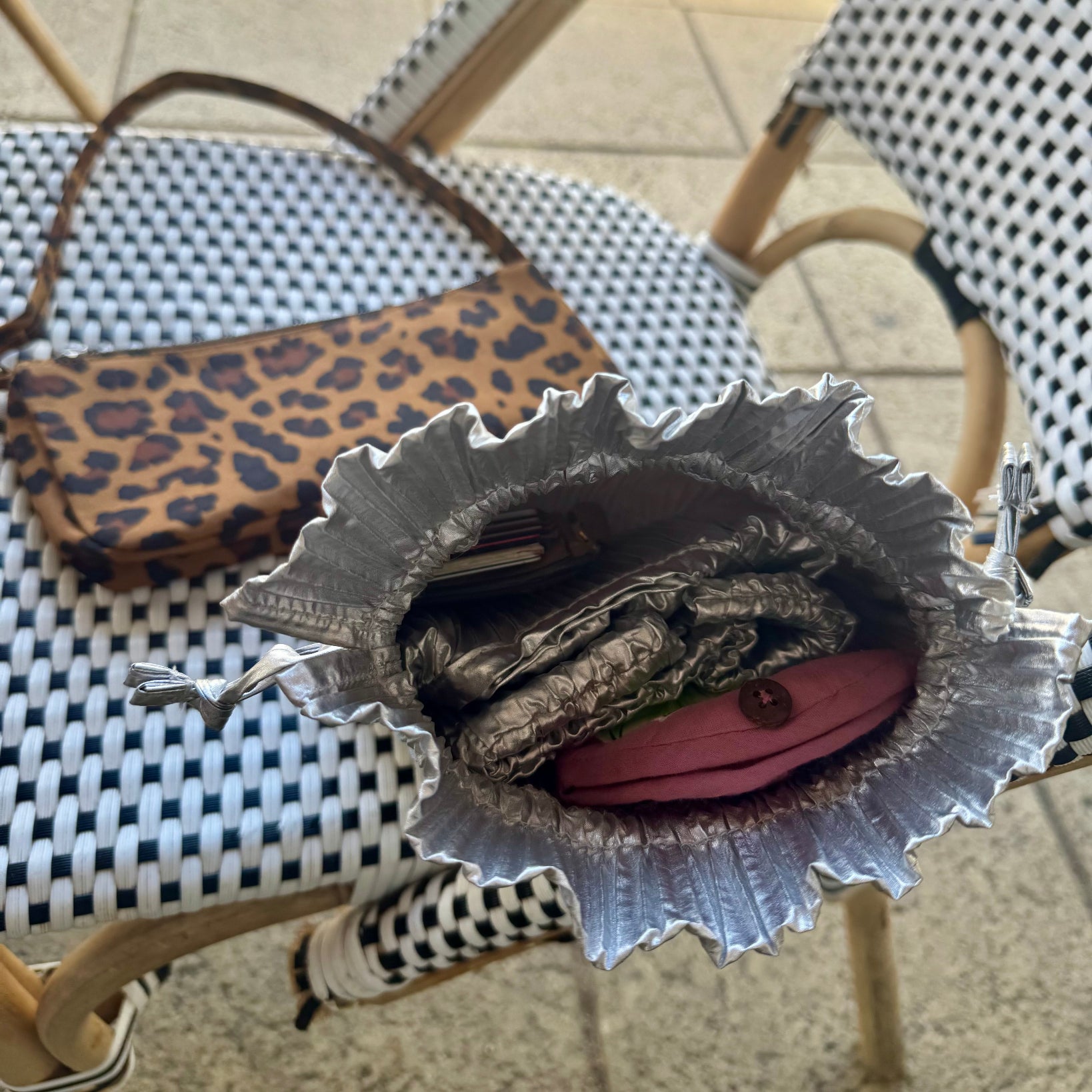 Inside of chrome metallic drawstring pouch on checkered chair with a leopard print clutch pochette outside.