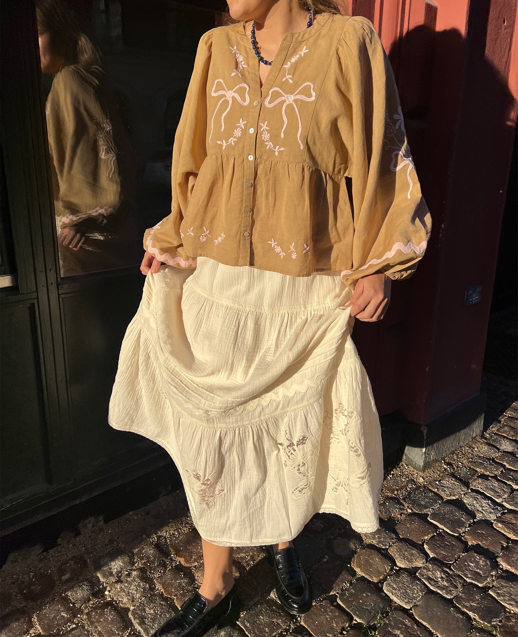 Woman wearing a light brown long sleeve top with button front and pink embroidery with a white skirt.
