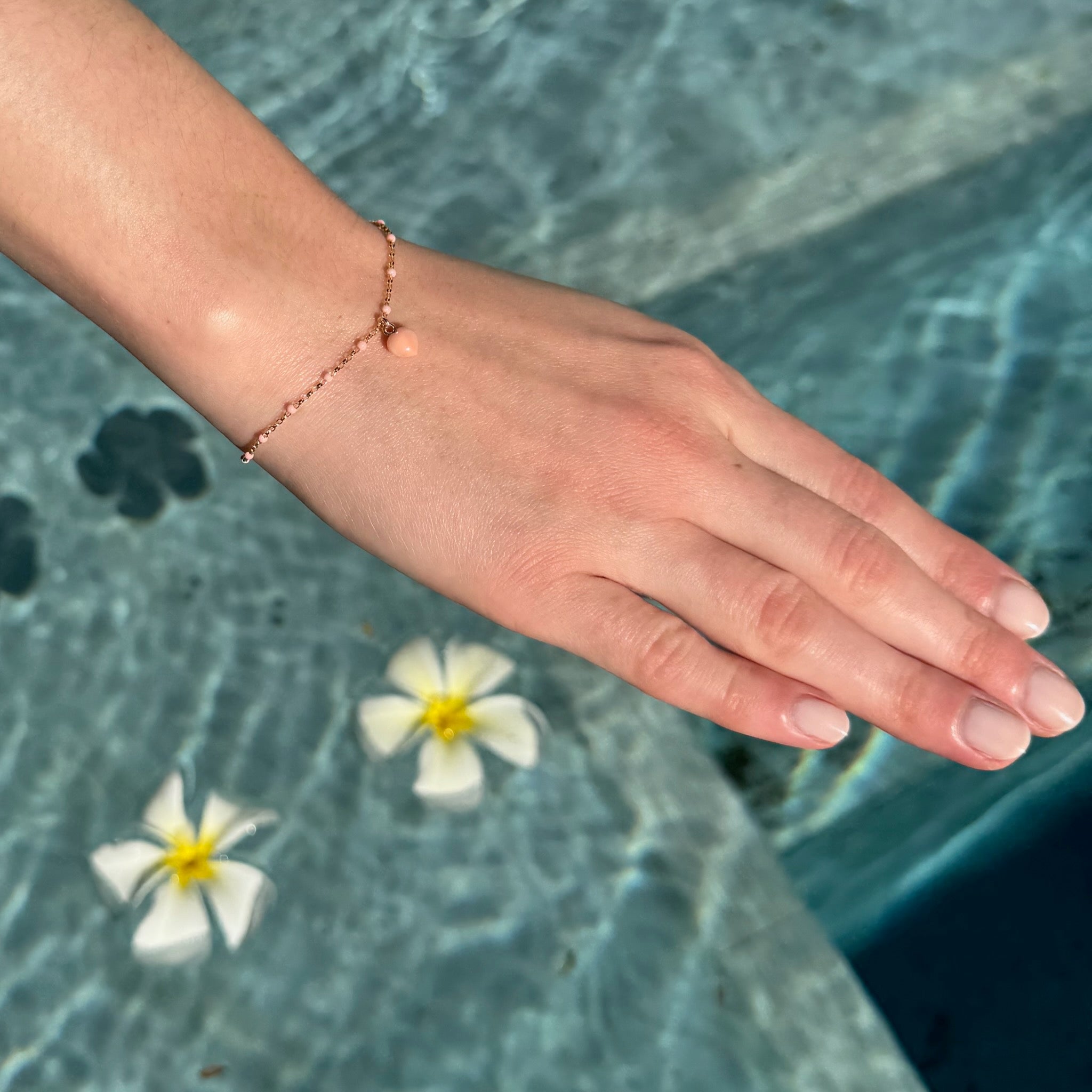 Coral Heart Bracelet in Seashell Pink on a woman's arm above a pool with flowers in water - ANNIE.G