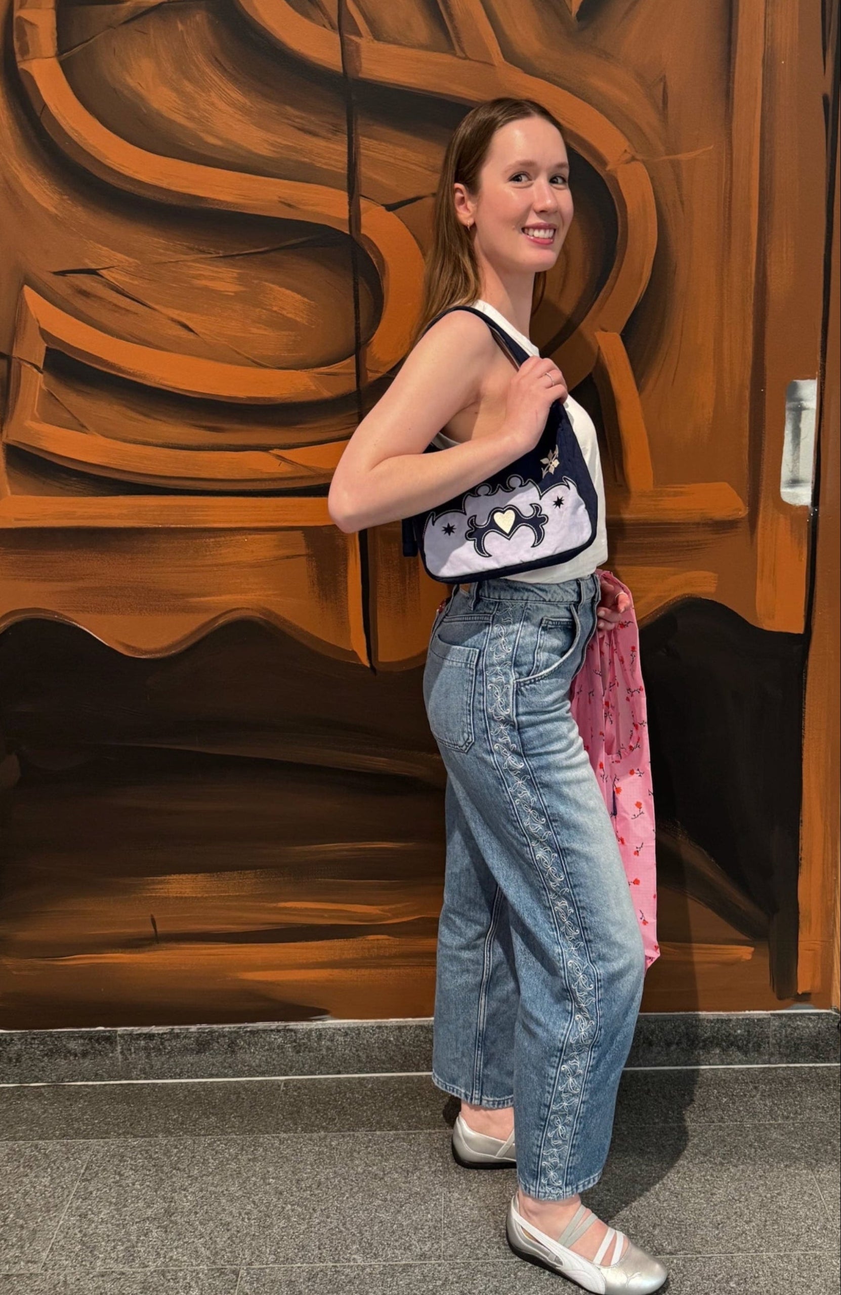 Woman wearing embroidered denim jeans with mini western style bag on shoulder and holding a pink shopping bag in front of a wooden door.