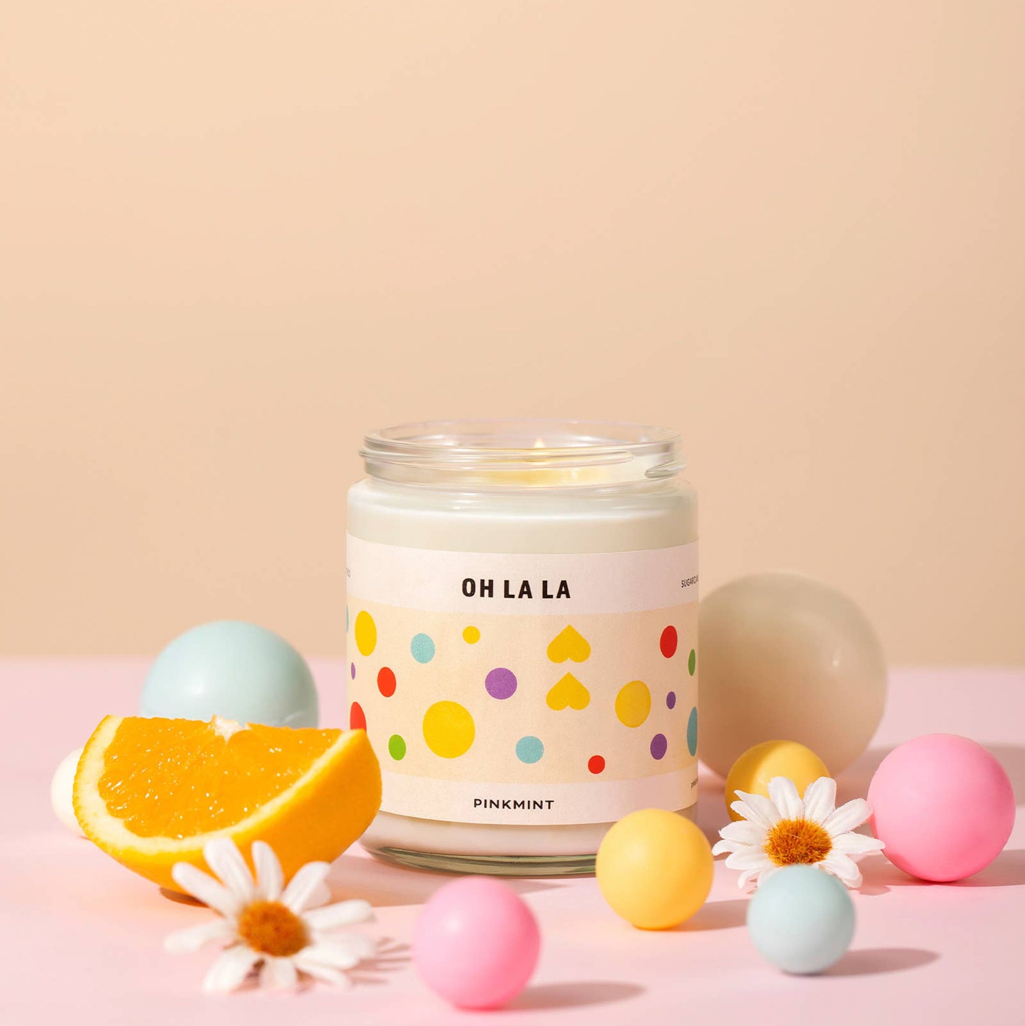 White soy candle in a jar with a wick lit and an orange sliced and flowers around it on a table - ANNIE.G