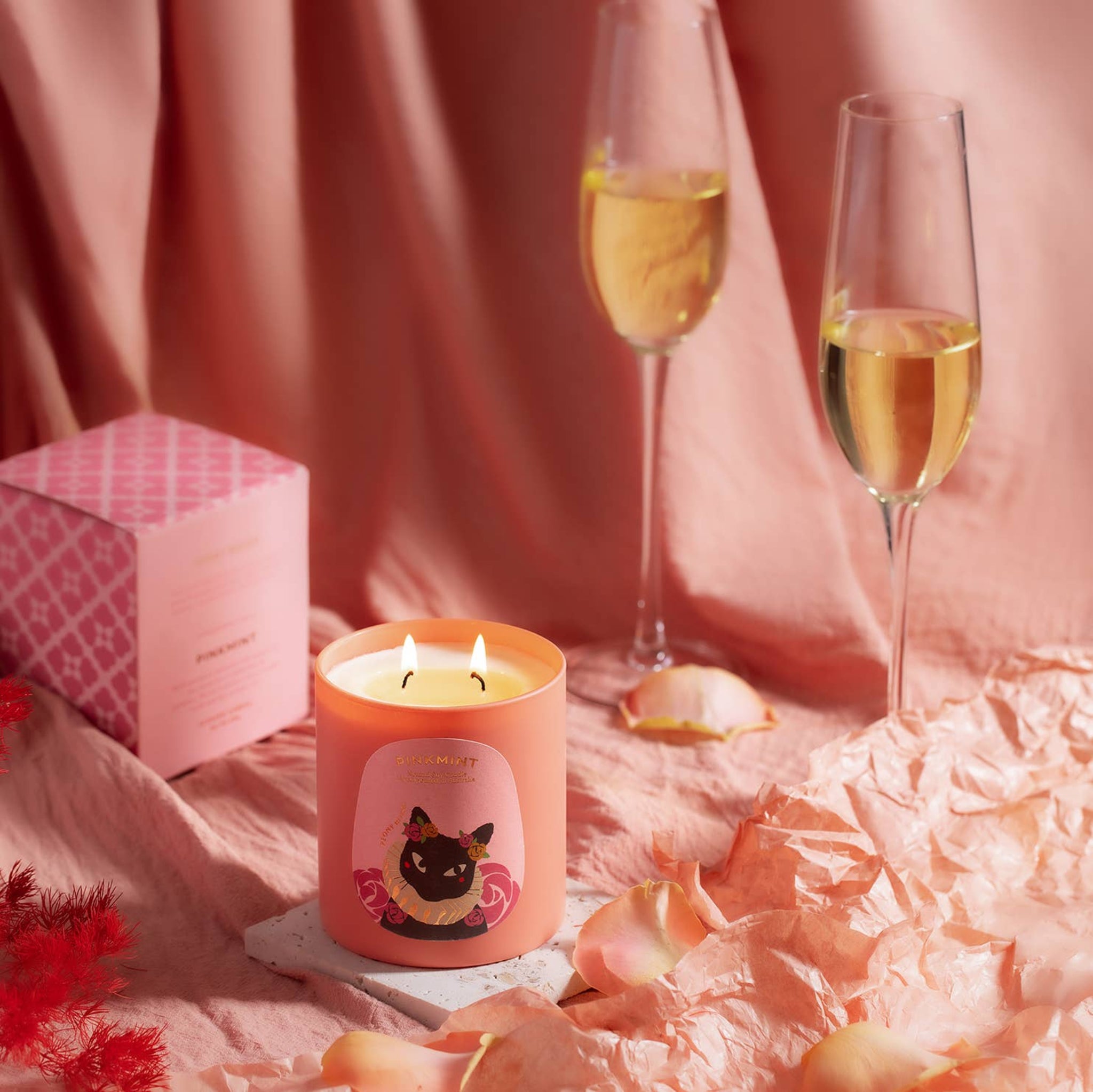 Pink candle jar with double wick lit and a pink presentation box on a tablecloth next to two champagne glasses - ANNIE.G