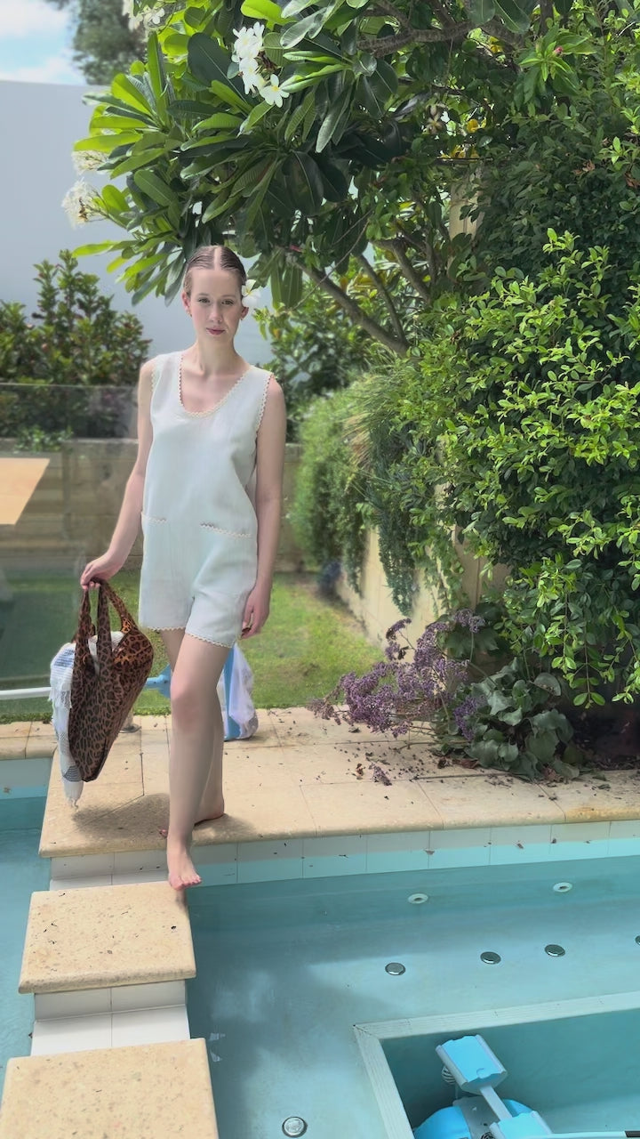 Woman in a light blue playsuit walking across pool pavers with a leopard bag in hand.