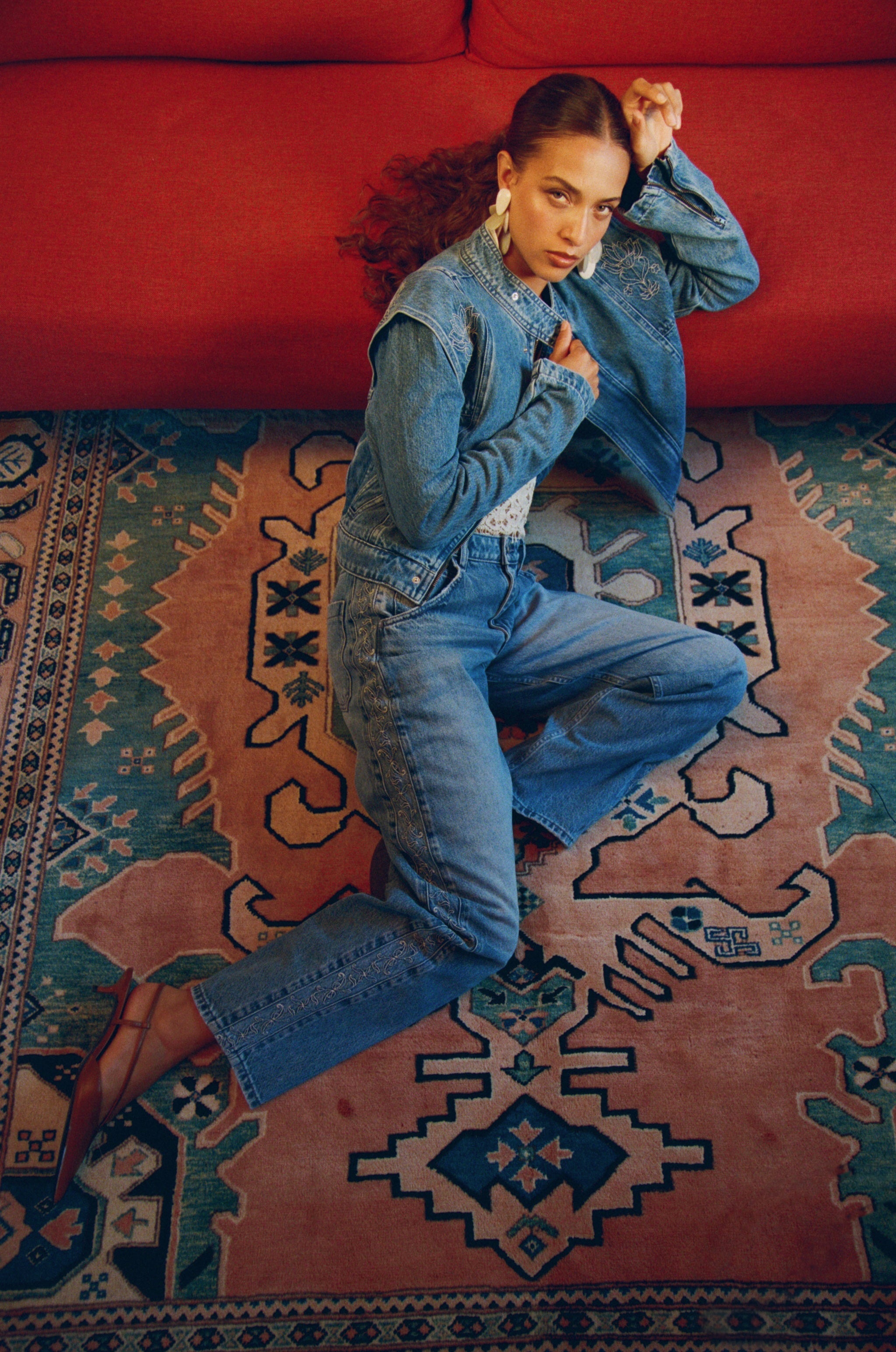 Woman in blue denim outfit sitting on a patterned rug with a red couch in the background.