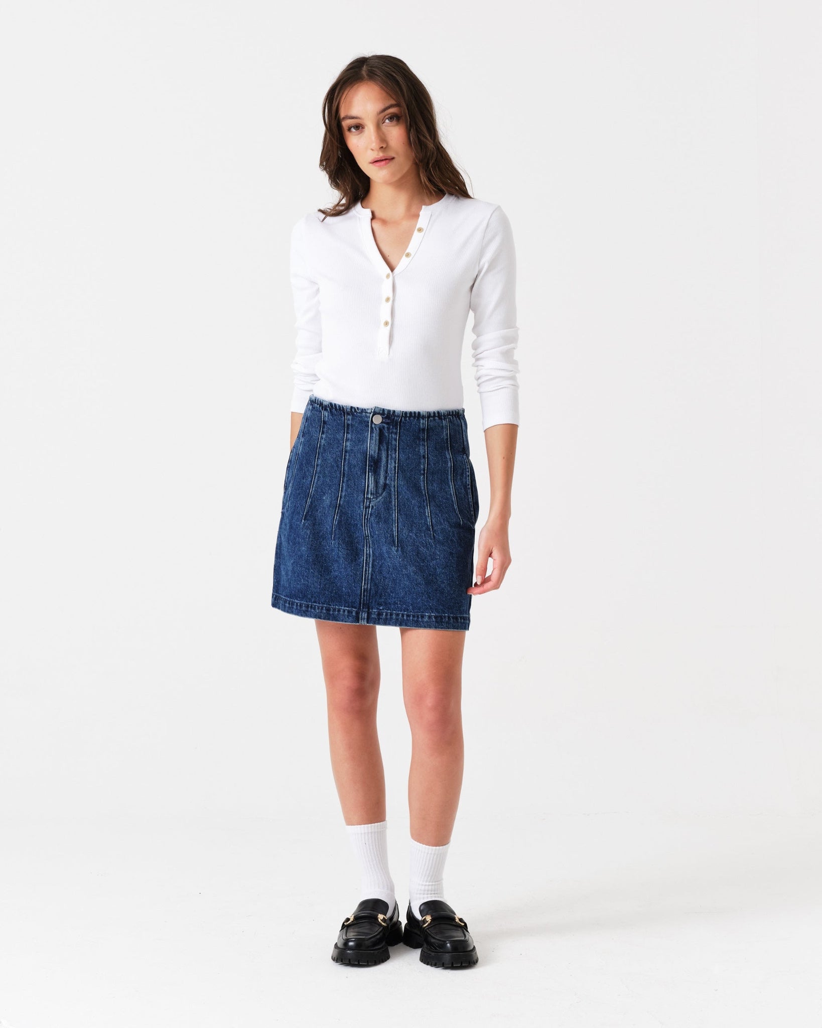 Woman wearing a white shirt, blue denim mini skirt with corset seam detailing in front.