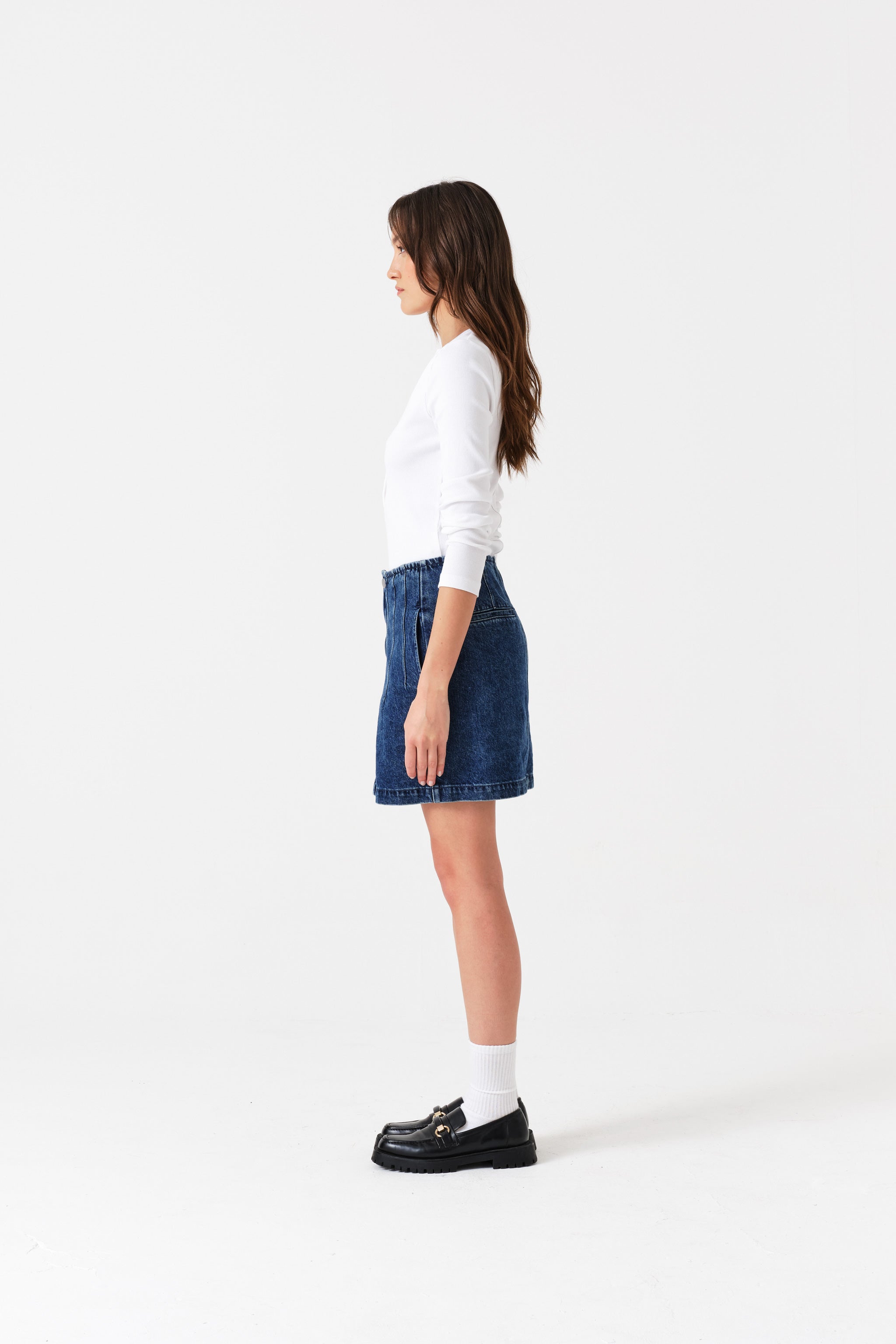 Person standing at the side and wearing a white sweater, blue mini denim skirt, and black shoes.