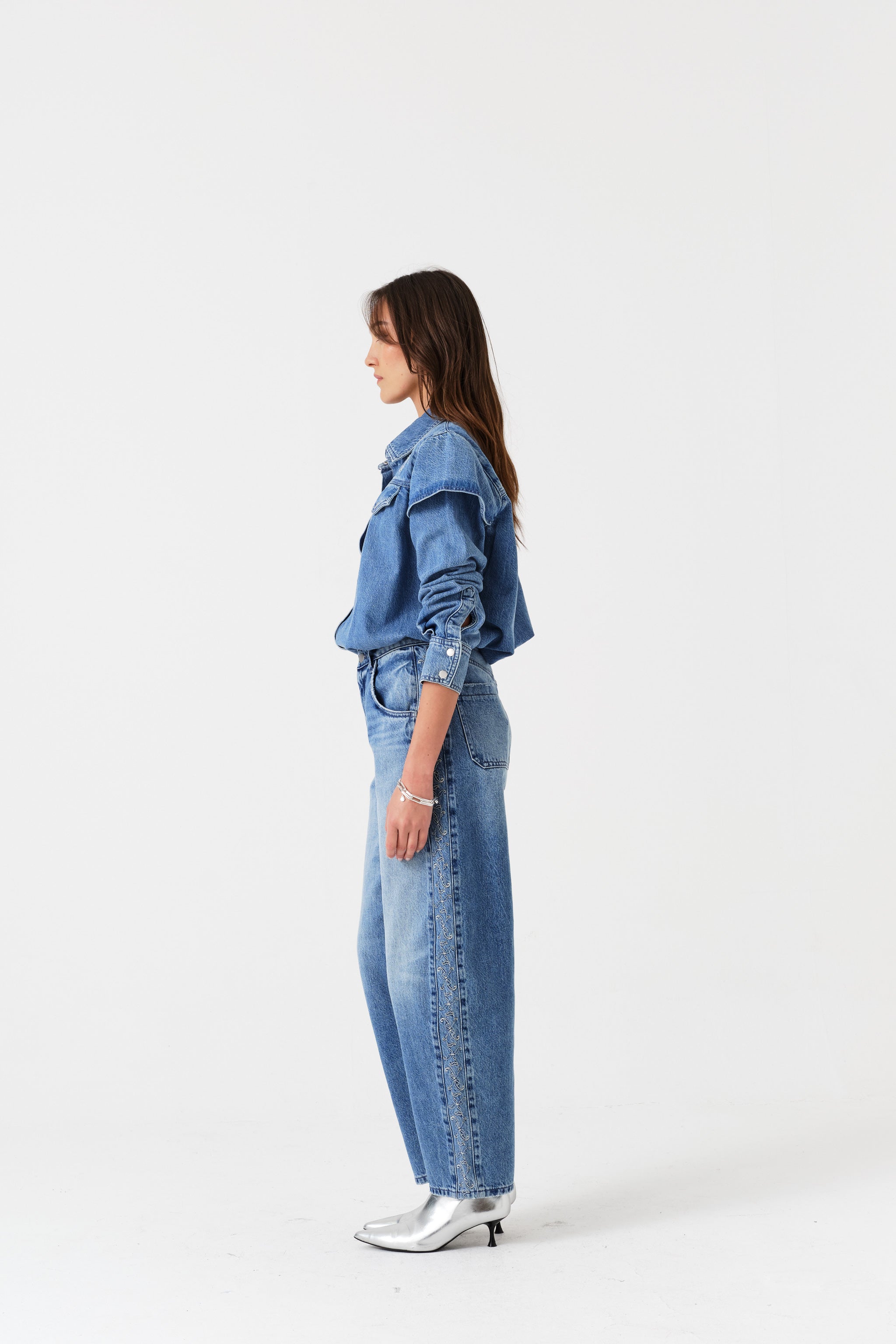 Woman standing at the side and wearing a denim blue shirt and embroidered jeans outfit.