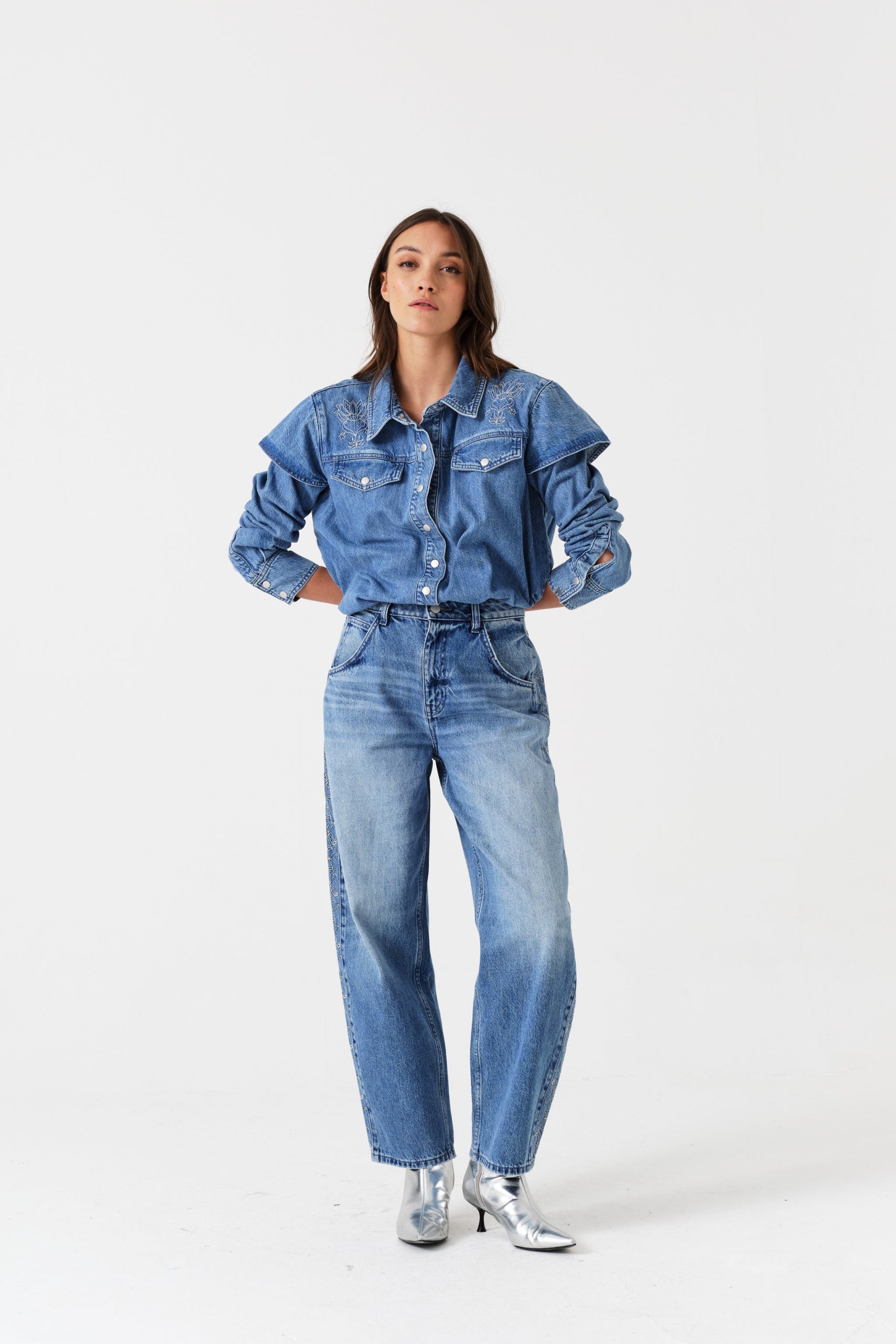 Woman wearing a denim blue shirt with embroidery and long sleeves and jeans with hands behind back.