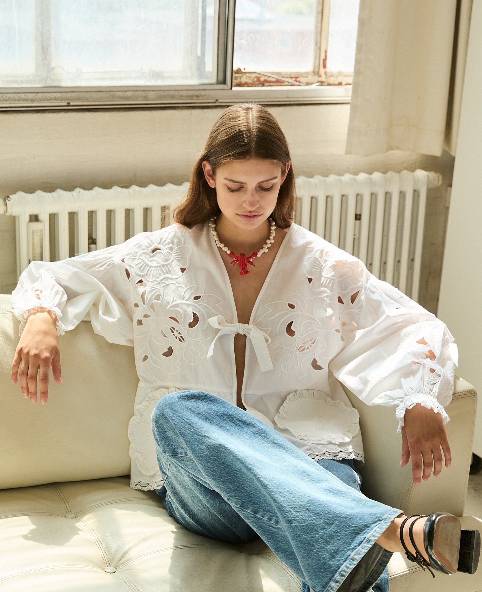 Woman sitting on lounge wearing a white top with long sleeves and front ties with cutwork embroidery, jeans and heels.