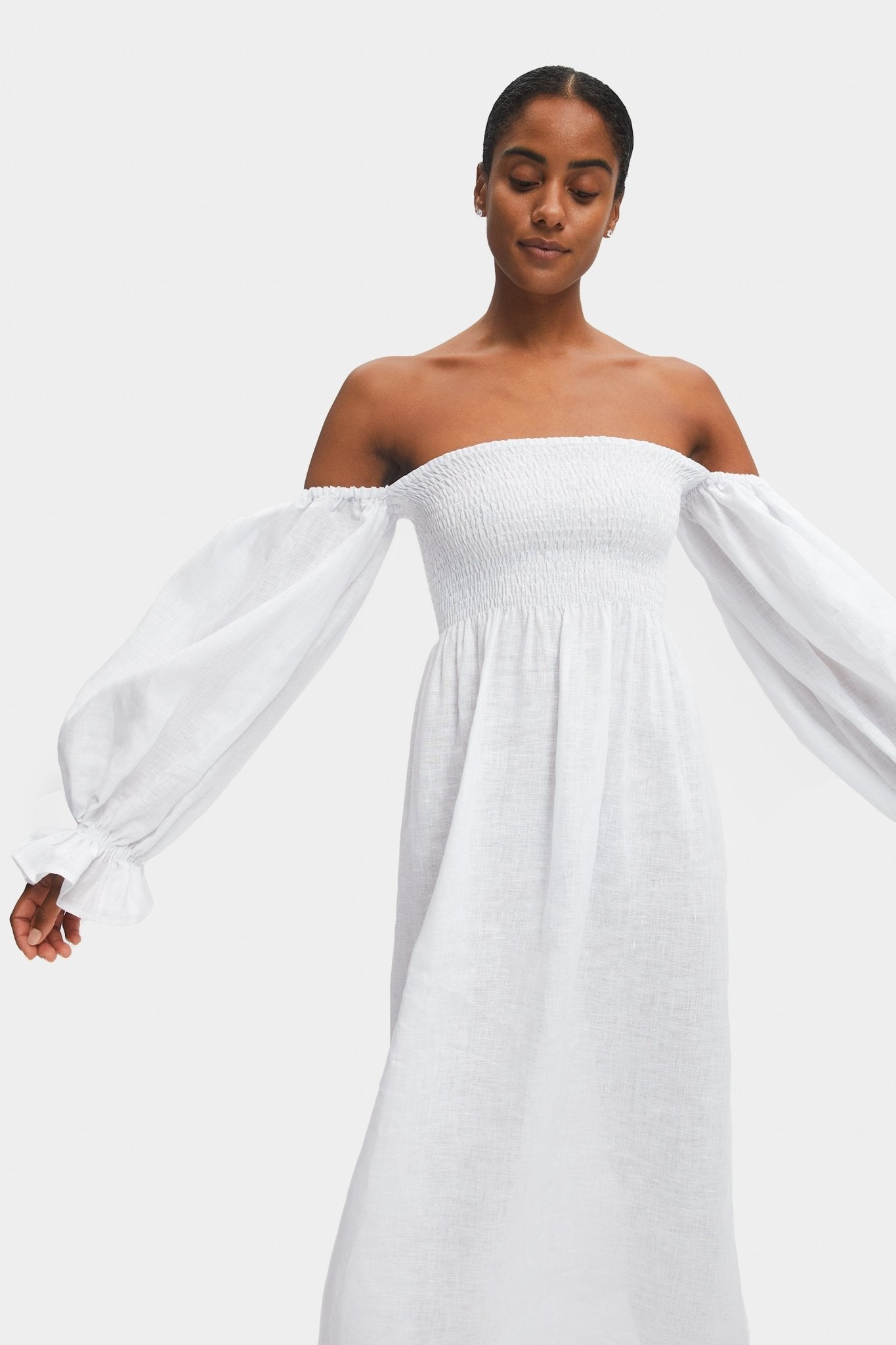 Woman with arms out to the side wearing a white off the shoulder dress with smocked bodice and long puffed sleeves with elastic cuffs and frill.