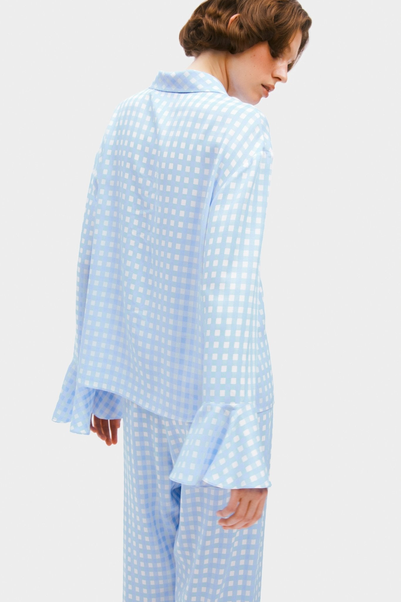 Woman wearing blue vichy pyjama set showing ruffles on long sleeve cuff and back of shirt with collar.