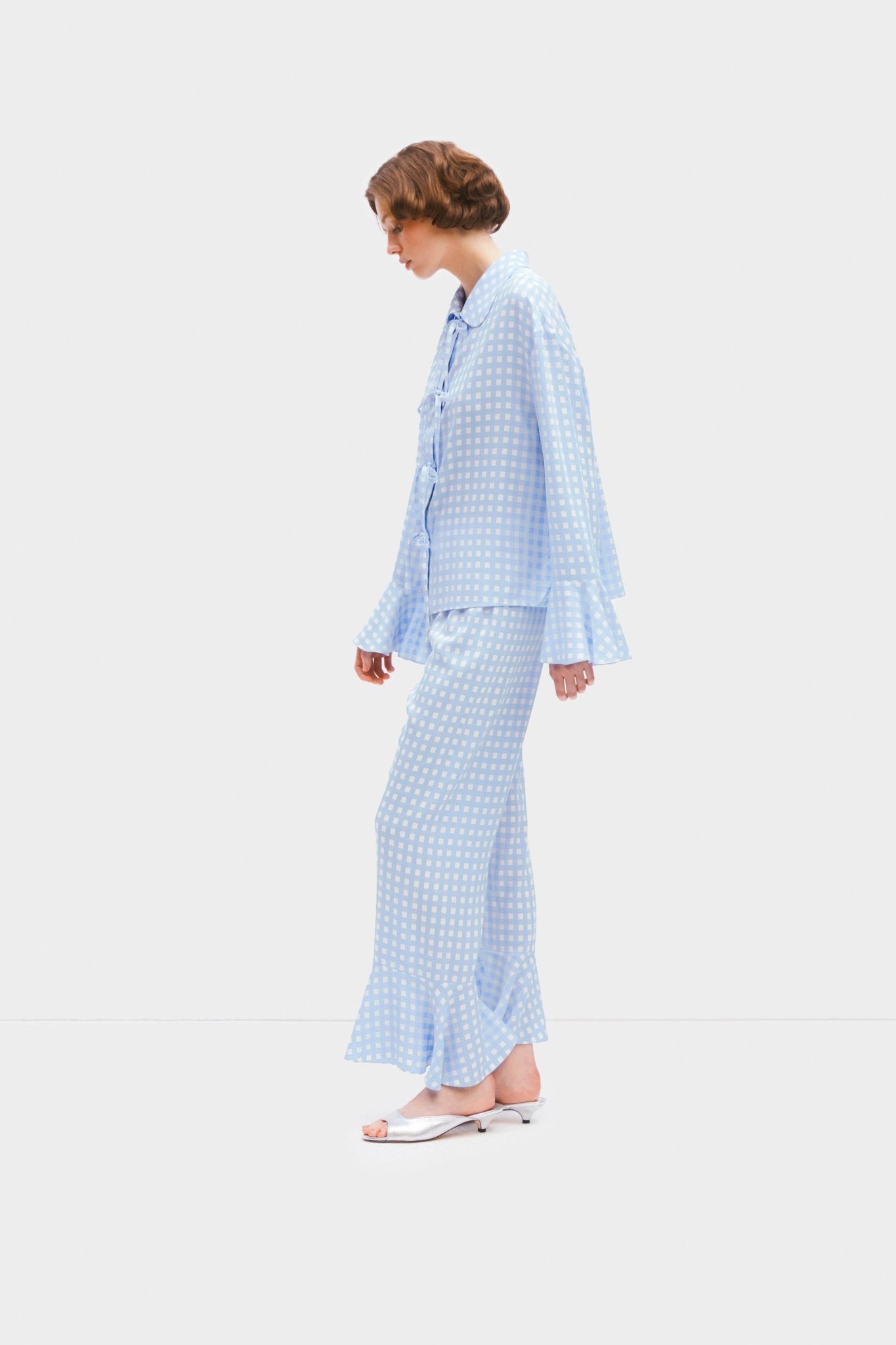 Side of a woman wearing blue vichy pyjama set of long sleeve shirt with ruffles at cuffs and pants with ruffles at leg opening.