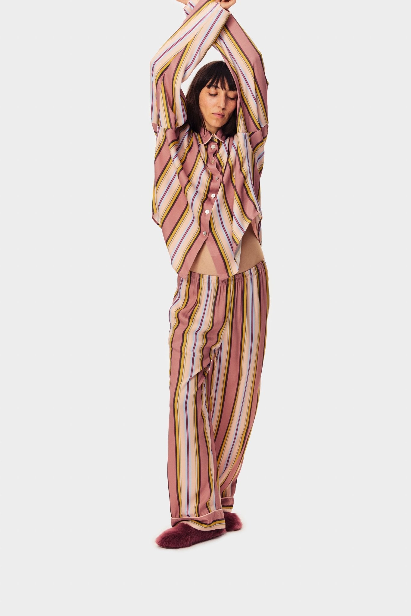 Woman wearing pink striped buttoned shirt and long pants with hands in the air showing elastic waistband.
