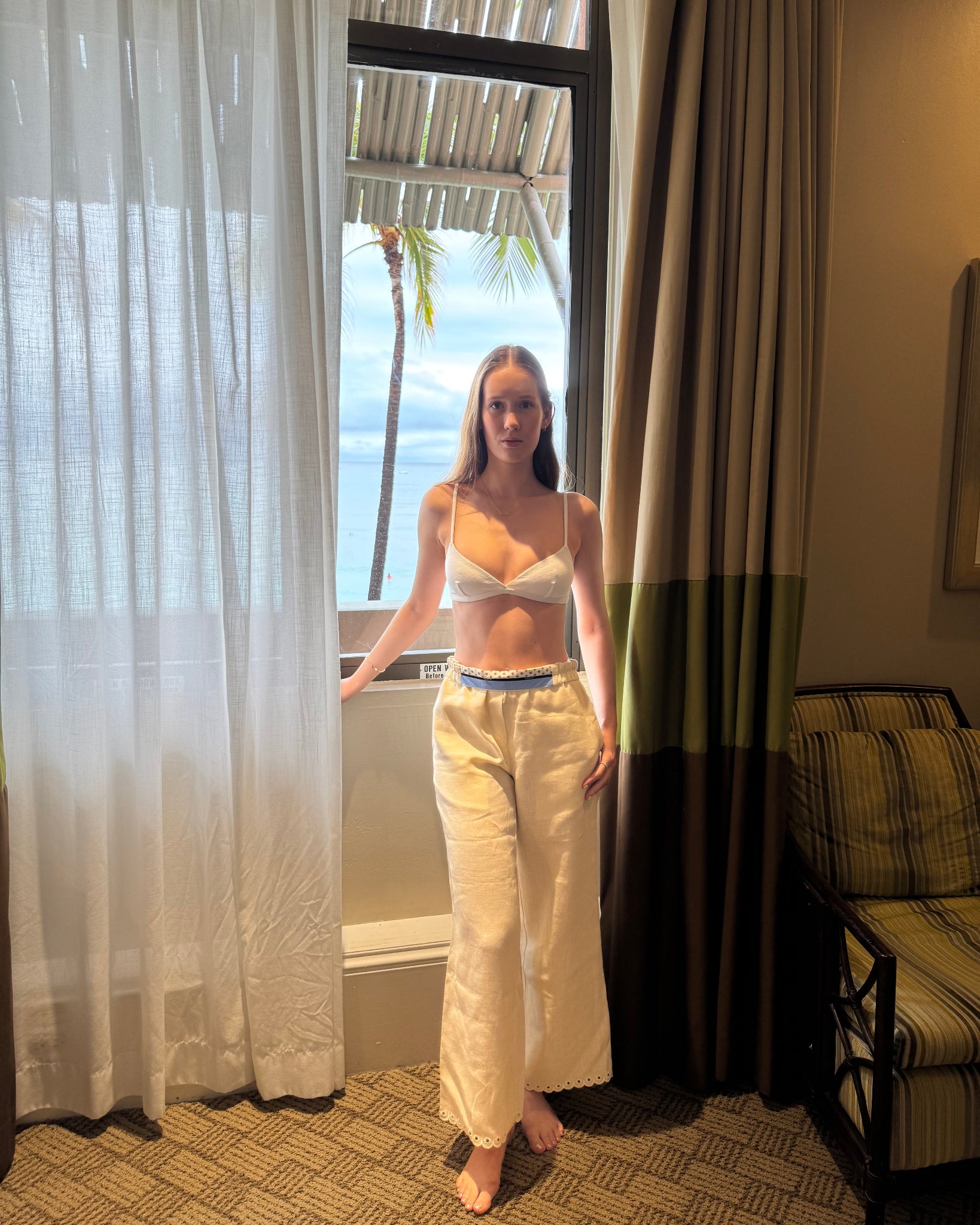 Woman wearing a bikini with cream linen long pants with lined waistband folded over and scalloped cropped hem standing in front of a window with a scenic view outside.
