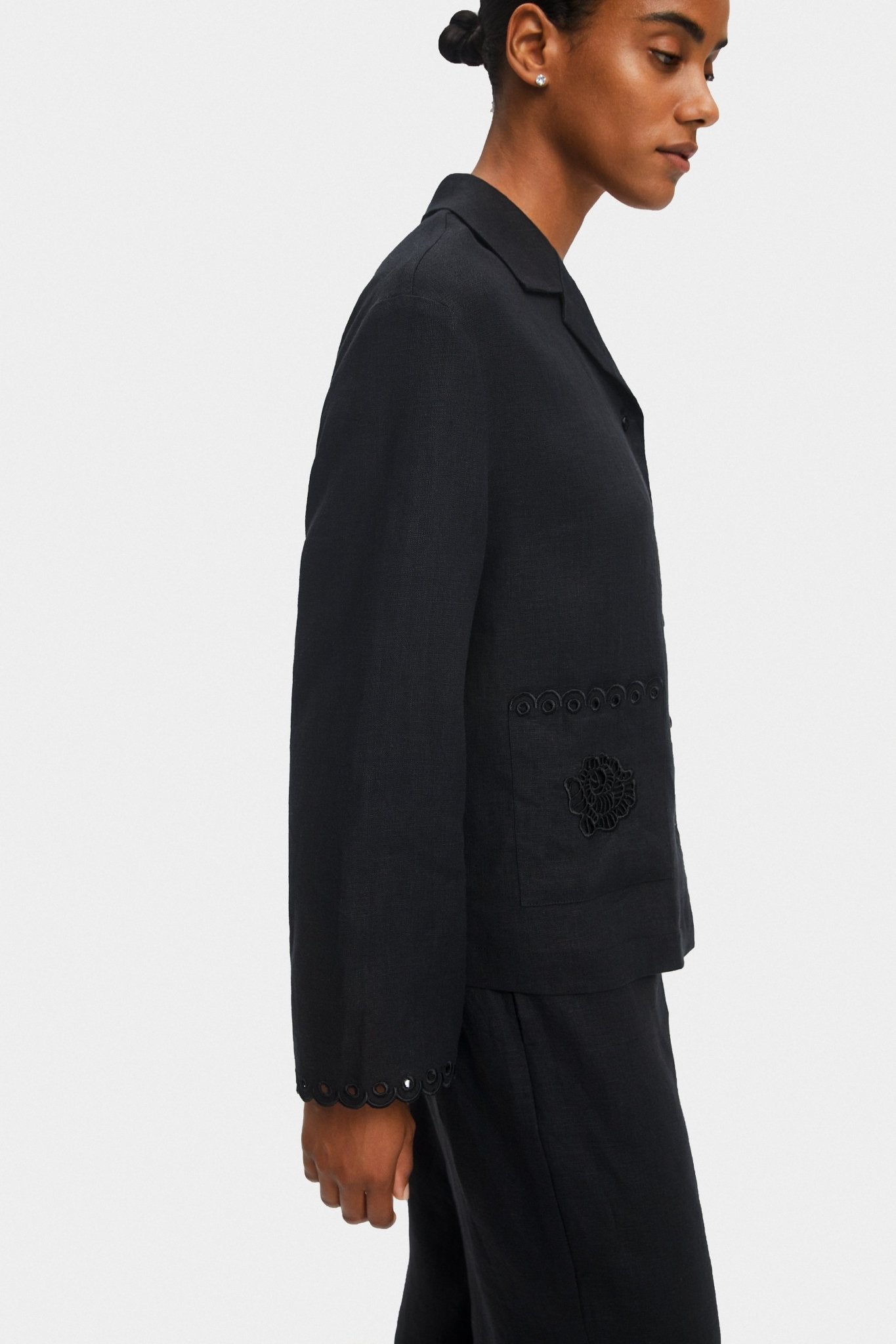 Woman standing on the side and wearing a black long sleeve shirt with scallop hem on sleeves and long pants.