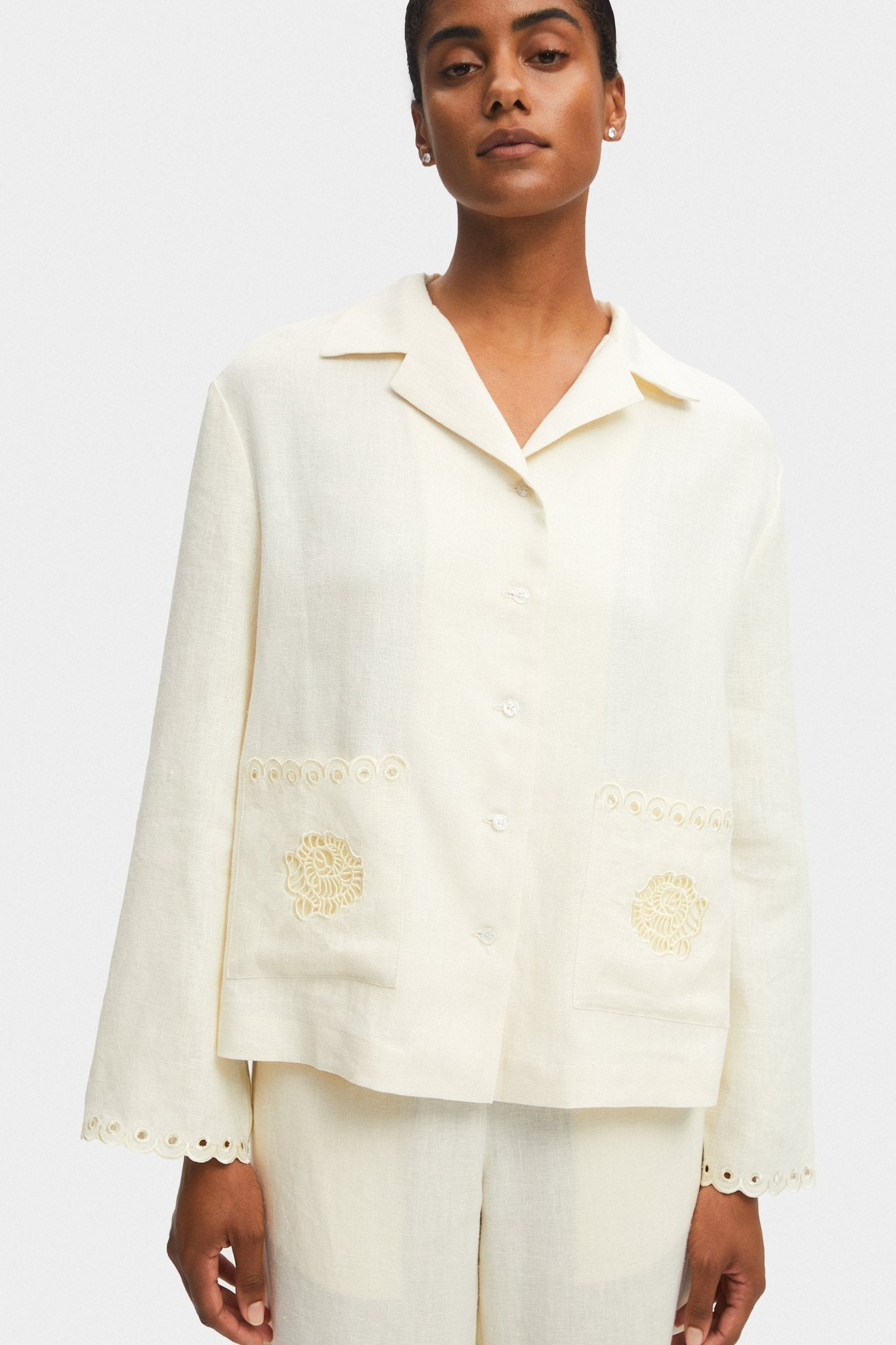 Woman wearing a cream long sleeve shirt with scallop hem on cuffs and embroidery on pockets with front buttons.
